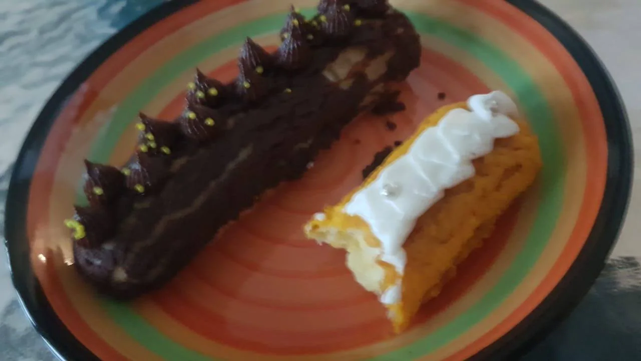 The image features a close-up view of two pastries on a colorful plate. The plate has concentric rings of orange, green, and black, adding a vibrant backdrop to the desserts. On the left is a chocolate pastry, adorned with dark chocolate icing piped in small peaks. These peaks are decorated with tiny yellow sprinkles, giving it a decorative touch. On the right, there is a pastry with a yellowish base, topped with white icing, arranged in a wavy pattern. The white icing is accented with small silver balls, providing an elegant contrast. The lighting in the image is soft and natural, highlighting the textures and colors of the pastries, and making the chocolate glaze appear rich and glossy. The setting appears to be a casual dining area, focusing on the appetizing presentation of these sweet treats.