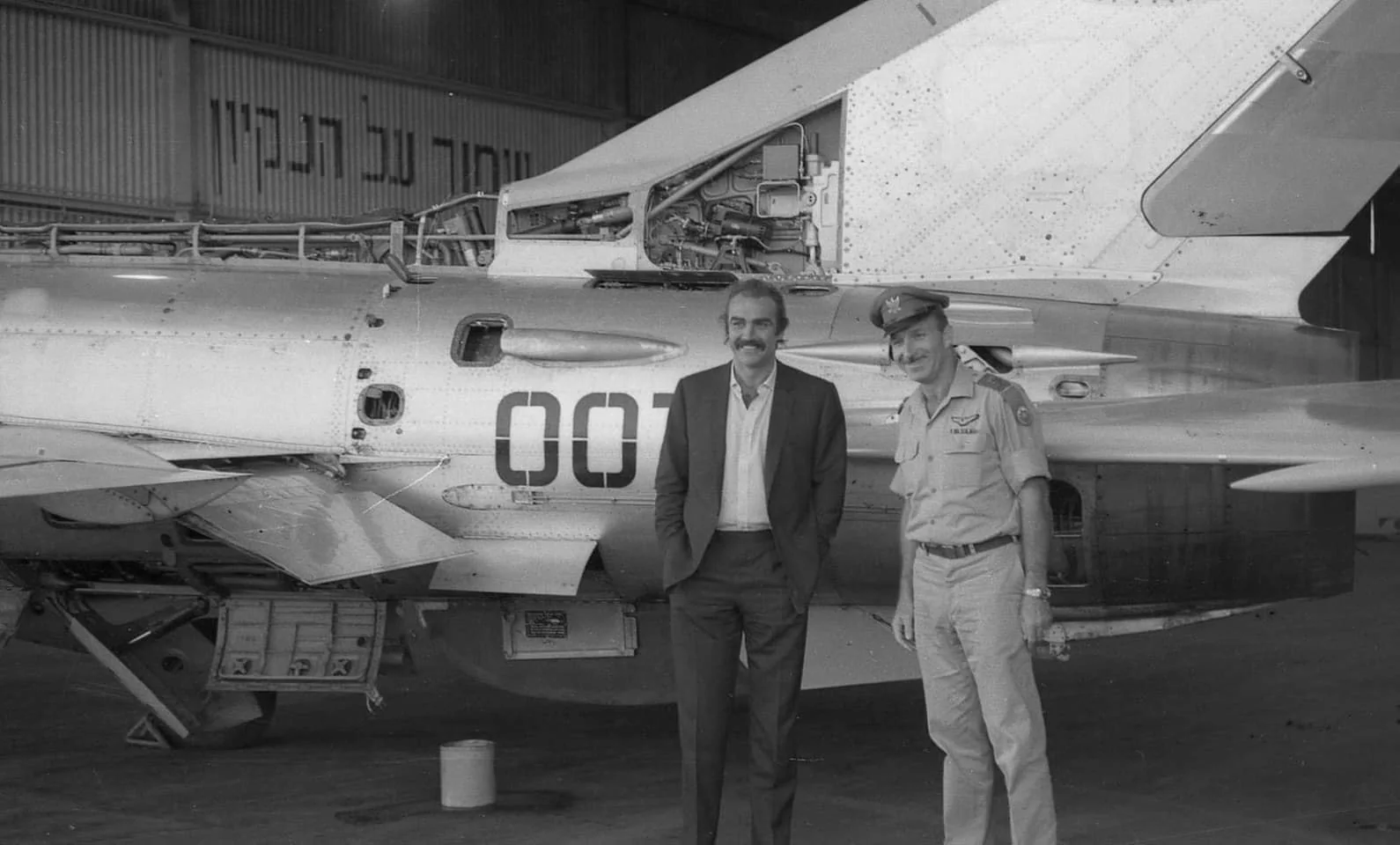 Sean Connery is seen standing next to an unidentified man in a military uniform inside a hangar. They are in front of a jet aircraft, which has the number "007" prominently displayed on its side, possibly referencing Connery's famous role as James Bond. The aircraft appears to be a Soviet MiG fighter, indicated by its design and features such as the wings and tail. The man beside Connery is wearing a uniform with insignia and a cap, suggesting he is a military officer. Connery is dressed in a dark suit with a light-colored shirt beneath. Both men are smiling and appear relaxed, suggesting a cordial or celebratory occasion. The background features a corrugated metal wall with Hebrew text, indicating the setting might be in Israel. The lighting is neutral, typical of an indoor environment, and the image is in black and white, giving it a historical feel.