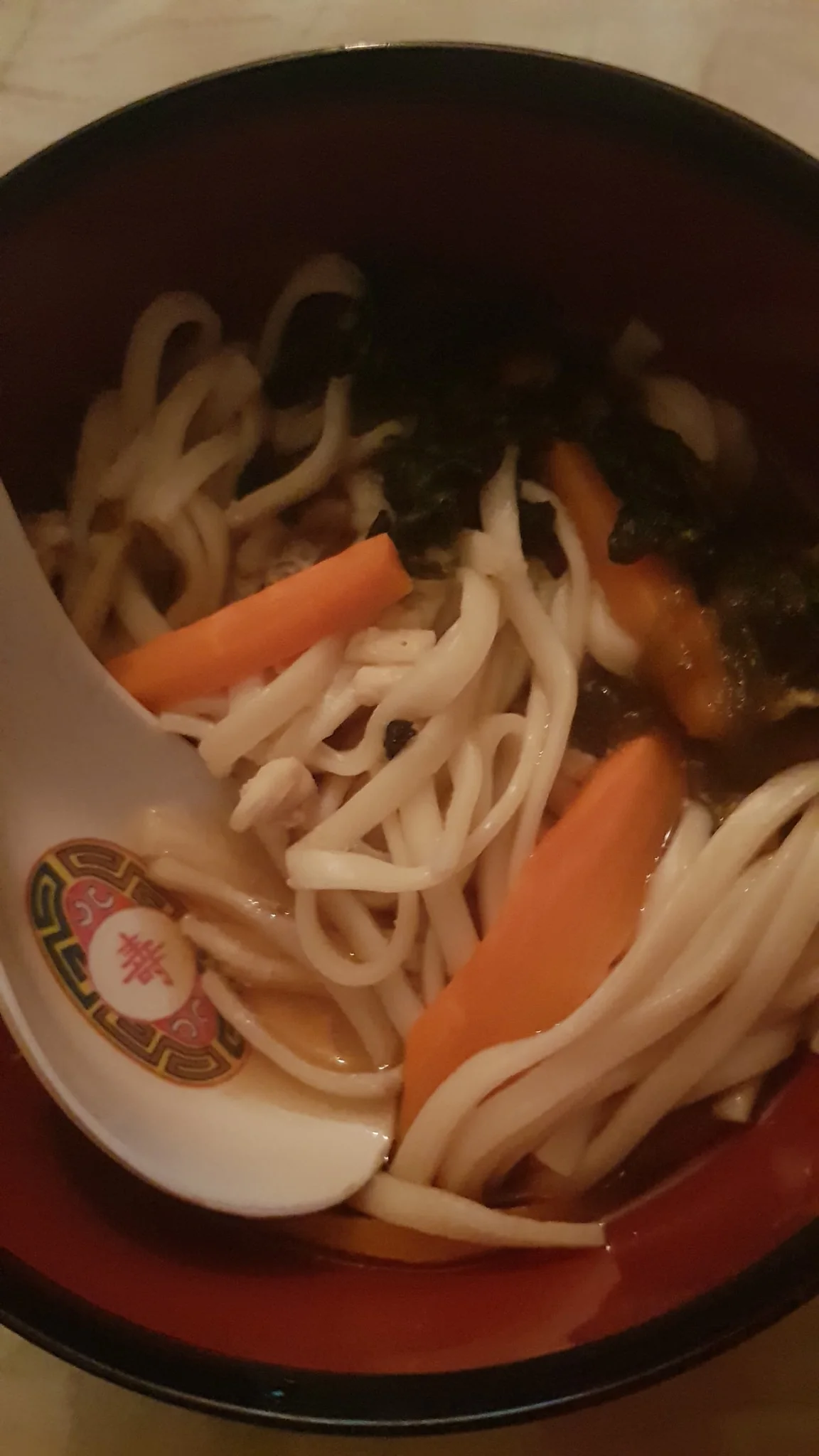 The image shows a bowl of ramen, featuring thick noodles, slices of carrot, and what appears to be leafy greens, all immersed in a flavorful broth. The bowl is red on the inside, and a decorative white ceramic spoon with Chinese-style motifs rests inside it. The noodles are prominently placed, with the carrot slices adding a splash of orange color to the composition. The lighting is warm and dim, suggesting a cozy indoor setting. This dish seems to be homemade, potentially part of a traditional Japanese meal. The caption hints that this ramen is made by "My mum," indicating a personal touch and familial connection. The post is likely from a setting where multiple people might be enjoying the meal together, as indicated by the mention of "all eight of my sister Lehara's friends."