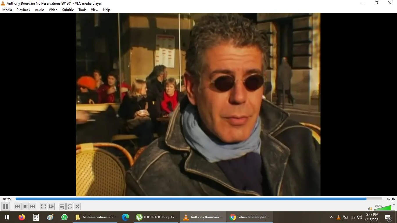 The image is a screenshot from "Anthony Bourdain No Reservations," specifically from the first episode of the first season. It features Anthony Bourdain wearing round sunglasses, a blue scarf, and a dark leather jacket. He appears to be sitting outdoors at a café, with wicker chairs visible in the foreground. The background shows several people sitting at tables, enjoying what looks like a sunny day. The setting suggests a casual and relaxed atmosphere, typical of a street café scene. The lighting is warm, indicating daytime, and the overall mood is leisurely, capturing the essence of enjoying a moment in a public space. The VLC media player's interface is visible, with a timestamp indicating about 40 minutes into the episode.