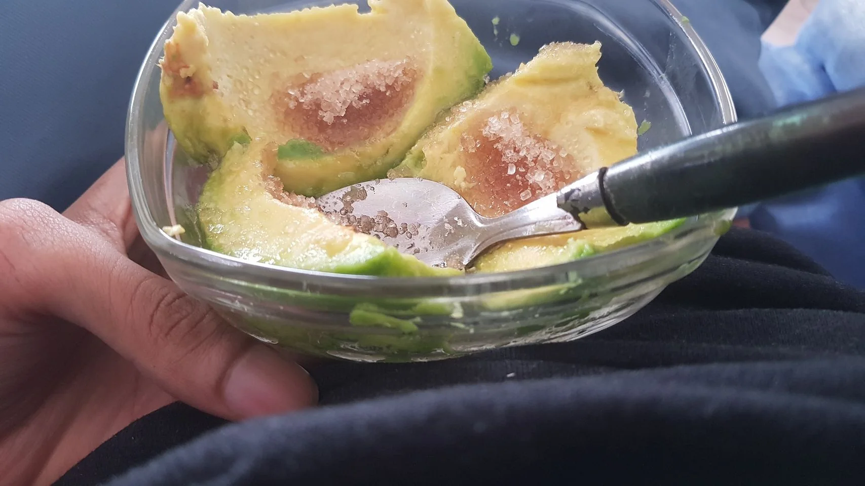 In this close-up image, a person is holding a small glass bowl in their hand. The bowl contains halved avocados, which are sprinkled with granulated sugar. A spoon rests inside the bowl, indicating that the avocados are being prepared for eating. The avocados have a rich green color with slightly brownish pits where the seeds were removed. The background is blurred, focusing attention on the bowl and its contents. The person is wearing a dark piece of clothing, which serves as a backdrop to the scene. The lighting is natural, emphasizing the texture of the avocado and the shine of the sugar crystals. The setting appears casual, perhaps at home, and the mood is relaxed, suggesting a simple, spontaneous snack.
