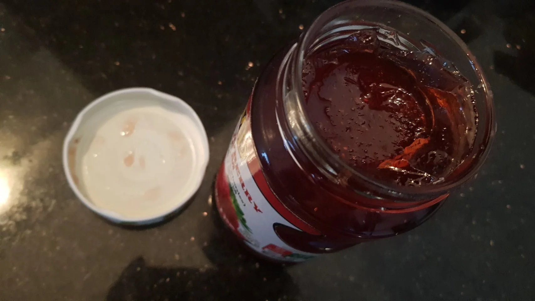 The image shows an open jar of raspberry jam placed on a dark countertop. The jar is filled with a deep red, glossy preserve, with some air bubbles visible on the surface, indicating it is freshly opened. The lid is placed beside the jar, showing remnants of jam on its underside. The label on the jar reads "Raspberry," suggesting the flavor of the jam. The scene is well-lit, allowing the deep red color of the jam to stand out against the dark background. There is no visible branding or other distinguishing text present, and the setting appears to be a kitchen countertop. The perspective is from above, focusing on the contents of the jar and the lid.