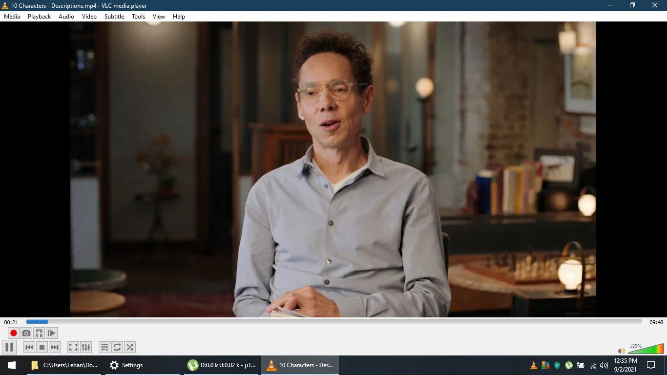 Malcolm Gladwell is featured in this screenshot from a video titled "10 Characters - Descriptions.mp4," playing in VLC media player. He is seated in an indoor setting, which appears to be a casual and possibly a study or library environment. Gladwell is wearing a light blue button-up shirt, and he is holding what seems to be a notebook or a book in his hands. He appears engaged in a conversation or interview, with a relaxed and thoughtful expression on his face.
The background is softly lit, with shelves and decorative items that suggest a cozy and intellectual ambiance. There is a wooden table with a chessboard, adding to the scholarly atmosphere. The video playback interface of VLC media player is visible at the bottom, showing standard controls and a timeline that is 21 seconds into a 9:48 long video. The screen displays the directory path “C:\\Users\\Lehan\\Documents,” indicating the file location, and network activity is shown as minimal ("D: 0.0 k U: 0.02 k - µTraffic"). The system clock reads 12:35 PM on 9/2/2021.
