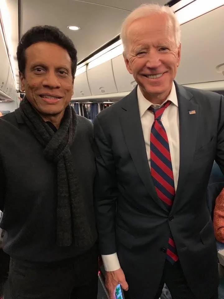 The image features Saroj Perera and Joe Biden inside what appears to be a train. Saroj Perera is on the left, dressed in a black sweater and scarf, smiling slightly. Joe Biden is on the right, wearing a dark suit with a white shirt and a red and blue striped tie. He is smiling broadly, holding a phone in his left hand. The setting suggests a casual encounter, perhaps during a journey. The background shows the interior of a train, with overhead compartments and soft lighting, creating a warm and friendly atmosphere. Both individuals are standing close together, suggesting a moment captured during travel. The overall mood appears cordial and pleasant, with both individuals seeming relaxed and at ease.
- Title: "..."
