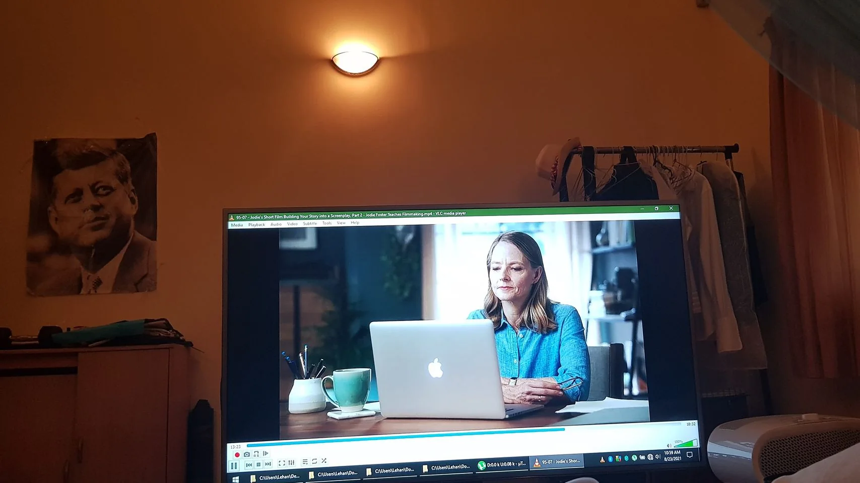 The image depicts a room with a television or computer monitor displaying a paused video. The video features Jodie Foster, who is sitting at a desk with a laptop open in front of her. She is wearing a blue shirt and appears focused on her work. The room has a warm, dimly lit ambiance, with a light fixture on the wall casting a soft glow.
On the screen, the VLC media player interface is visible, with menu options like "Media," "Playback," "Audio," "Video," "Subtitle," "Tools," "View," and "Help" at the top. The video file named "Jodie Foster Teaches Filmmaking.mp4" suggests that it is part of a series on filmmaking, specifically about building a screenplay. In the background of the room, there is a black and white poster of John F. Kennedy on the wall. To the side, there's a clothing rack with various garments hanging, adding to the personal and informal setting.