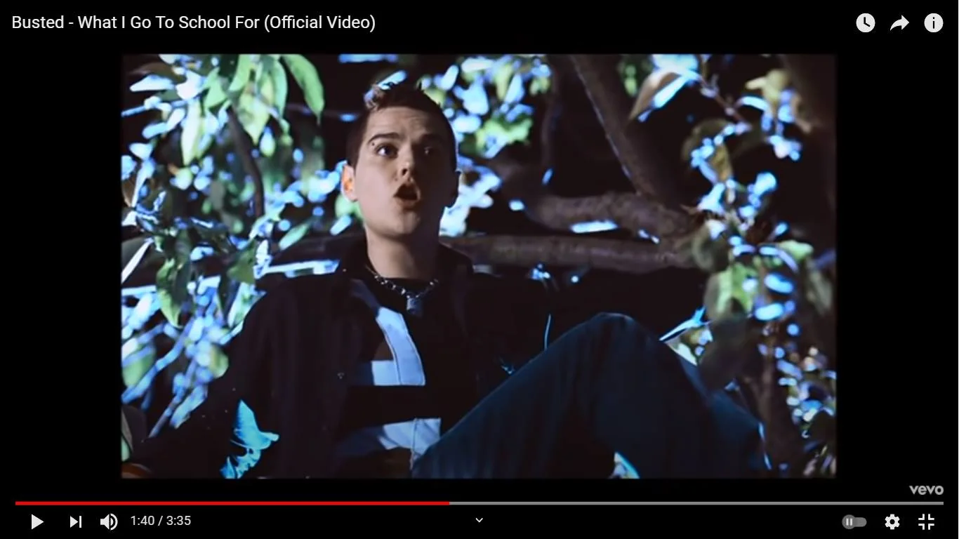 The image is a frame from the official music video for "What I Go To School For" by Busted. It shows a nighttime scene where a person is sitting in a tree. The lighting creates a dramatic contrast, highlighting the person against the dark background. The foliage surrounding the individual is lit, emphasizing the leaves and branches. The person is wearing a striped shirt under a dark jacket, with a chain necklace visible around the neck. Their mouth is open, suggesting they might be singing or speaking. The overall mood is energetic and rebellious, fitting the theme of the song. The video is published by Vevo on YouTube. The scene captures a youthful and adventurous spirit, consistent with the song's lyrics and playful tone.
- Title: "Busted - What I Go To School For (Official Video)"
- Site / app: "YouTube"
- Publisher: "Vevo"