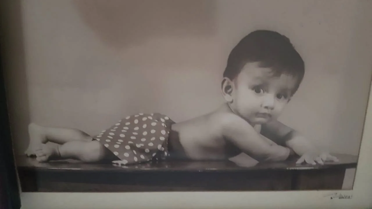 The image is a vintage black-and-white portrait of a baby identified as Lehan Edirisinghe. The baby is lying on their stomach, looking directly at the camera with a curious expression. They are positioned on a flat surface, possibly a table, covered with a piece of fabric with polka dots. The setting appears to be a studio, suggested by the smooth background and professional composition. The baby’s hair is neatly styled, and their eyes are wide open, adding a charming and innocent allure to the photograph. The soft lighting accentuates the baby’s delicate features and the texture of the fabric. The overall mood is sweet and timeless, capturing a precious moment in infancy. There is a signature or text in the lower right corner, which might be the photographer's mark.