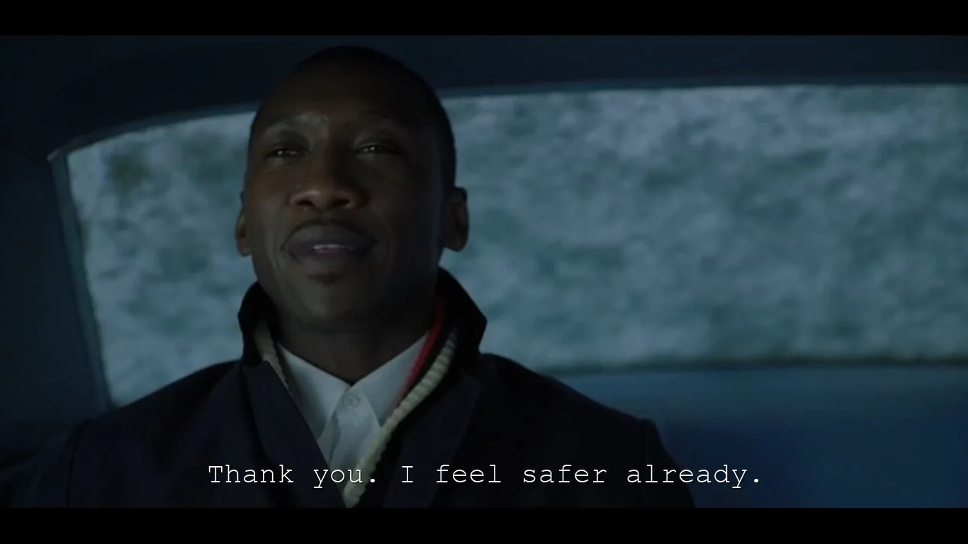 The image is a video frame showing a person sitting inside a car. The individual is wearing a dark coat over a white collared shirt, with a sweater visible underneath, indicating a possibly cold setting outside. The background suggests a snowy environment seen through the car window, enhancing the sense of winter or a journey through a cold region. The person appears to be speaking, as indicated by the subtitles at the bottom of the frame which read, "Thank you. I feel safer already." The lighting inside the car is dim, creating a cozy yet serious atmosphere. The expression on the individual's face suggests calmness or perhaps subtle gratitude. This frame likely captures a moment of dialogue or reassurance within a narrative, though the specific context or identities are not discernible from the image alone.