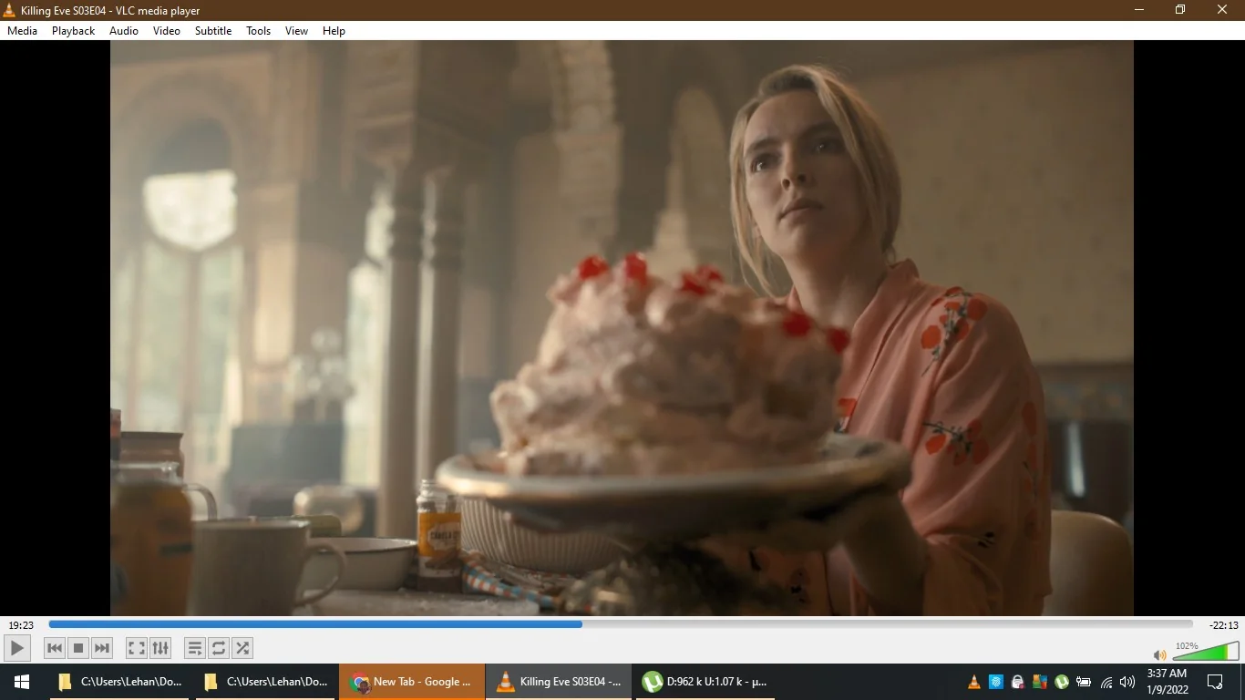 The image is a frame from the TV show "Killing Eve," specifically from Season 3, Episode 4. It is being played on VLC media player, as indicated by the visible player interface. The scene features Jodie Comer, who is holding a large plate with a cake covered in whipped cream and cherries. She is wearing a pink robe with a floral pattern and has a serious or contemplative expression on her face.
The setting appears to be a kitchen or dining area with a warm, softly lit ambiance, suggesting daytime. In the background, there are tall arched windows allowing natural light to flood the room, along with decorative elements that give the room an elegant look. A coffee mug and a jar of marmalade are visible on the table, adding to the cozy, domestic atmosphere. The video is paused at 19:23, with a total length of 22:13, as seen in the VLC interface.