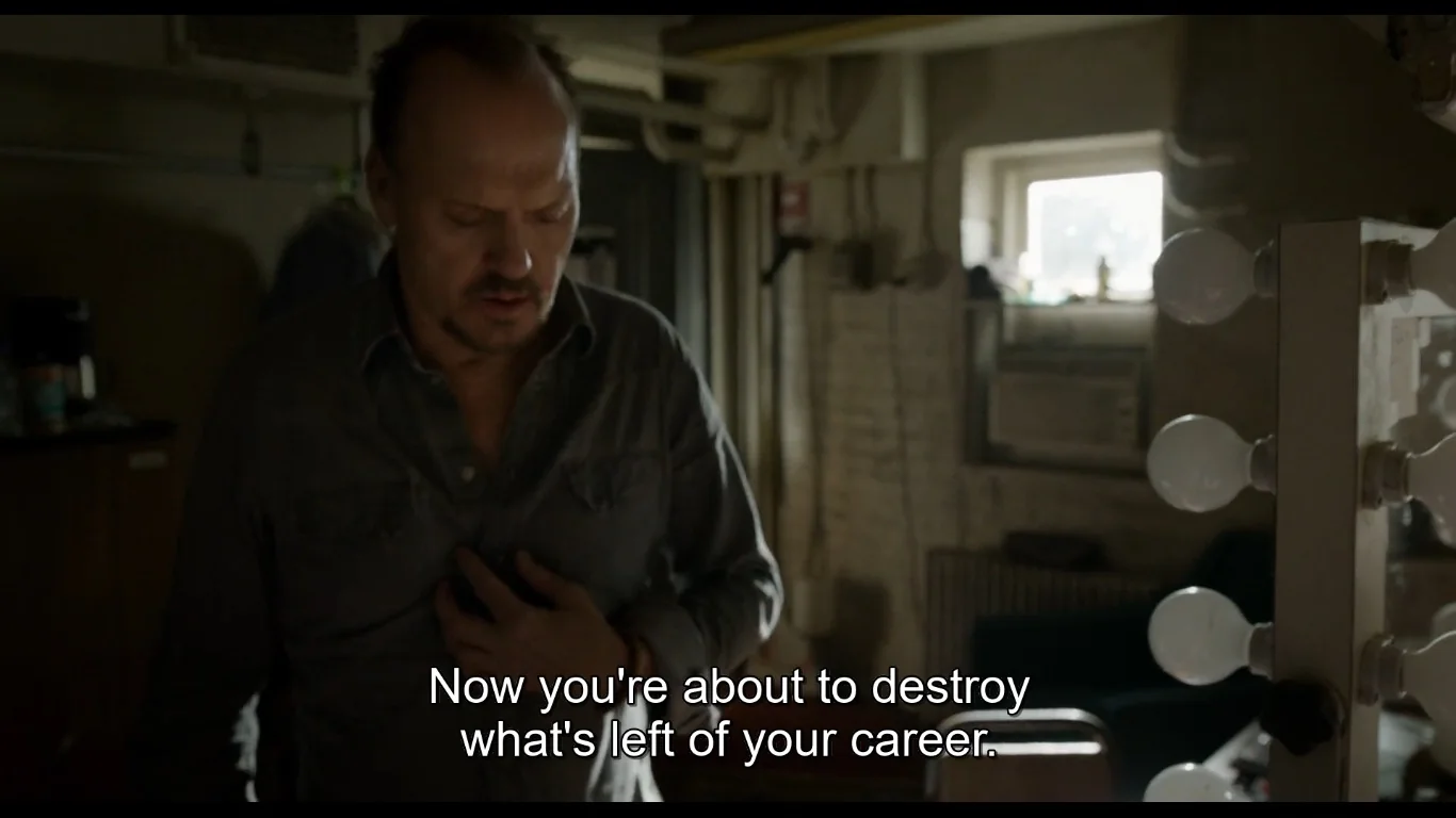 In this scene from the film "Birdman," Michael Keaton's character is captured in a tense moment inside what appears to be a dimly lit dressing room. The lighting casts shadows, creating a somber atmosphere that matches the serious tone of the situation. Keaton, wearing a dark gray button-up shirt, appears deep in thought, his hand placed on his chest as if contemplating or feeling stressed. The room is cluttered, with a makeup mirror lined with light bulbs visible on the right side of the frame, suggesting a theater setting. In the background, there is an air conditioning unit under a small window, adding to the room's utilitarian feel. The subtitle reads, "Now you're about to destroy what's left of your career," indicating a critical and introspective moment for the character. The entire composition and mood reflect themes of self-reflection and the pressures of maintaining a career in the arts.