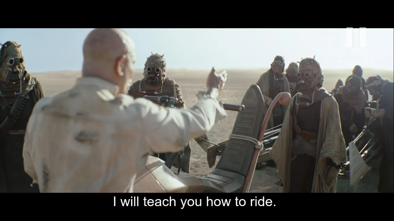 In this video frame, a group of characters is gathered in a desert landscape, creating a sense of an expansive, arid environment. The central figure, seen from behind, is speaking to a group of masked individuals. These individuals wear distinct, rugged outfits typical of desert nomads, with layered robes and goggles, suggesting they belong to a unique tribe or group accustomed to harsh conditions. The lighting is bright, indicative of daylight in a desert setting, enhancing the isolation and vastness of the location.
In the foreground, a vehicle with a vertical handlebar and a padded seat is partially visible, hinting at its use for transportation across the sandy terrain. The speaker, dressed in light-colored clothing, extends an arm toward the group, indicating an offer or an instruction. The caption at the bottom reads, "I will teach you how to ride," suggesting a moment of learning or alliance-building between the characters. The scene conveys themes of survival, mentorship, and collaboration within a challenging environment.