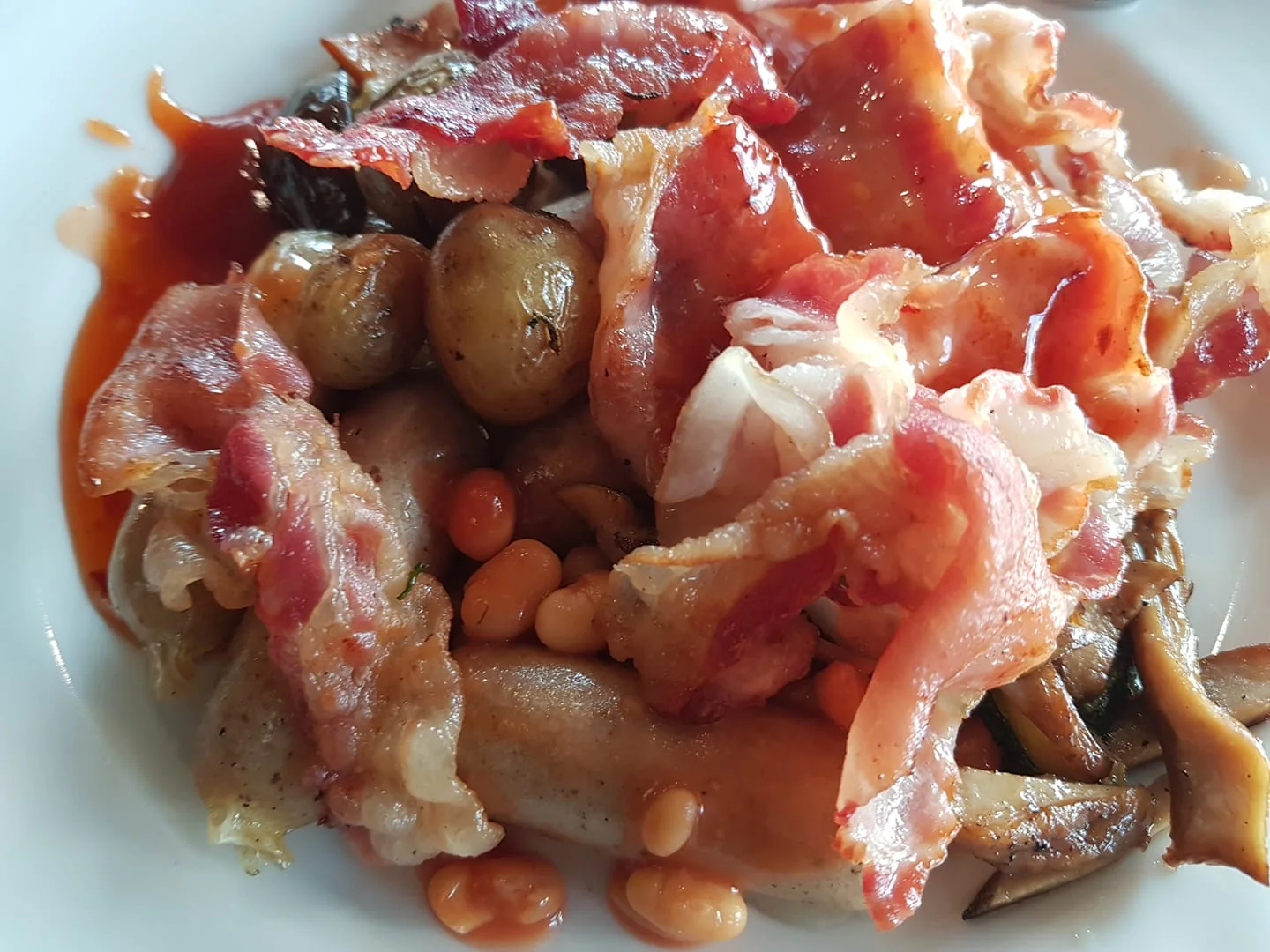 This image showcases a hearty plate of breakfast food, featuring several classic elements. In the foreground, crispy fried bacon strips are layered generously across the dish, providing a rich, savory appeal. Nestled among the bacon are golden-brown potatoes, which appear to be roasted, adding a rustic texture. Below the bacon and potatoes, baked beans are visible, offering a hint of sweetness and a pop of orange color. A sausage peeks out from beneath the layers, contributing to the plate's substantial, protein-rich composition. Off to the side, sautéed mushrooms add an earthy flavor to the ensemble. The colors are warm and inviting, dominated by reds, browns, and golden hues, typical of a traditional English breakfast. The overall presentation is casual yet appetizing, evoking a sense of comfort and indulgence.