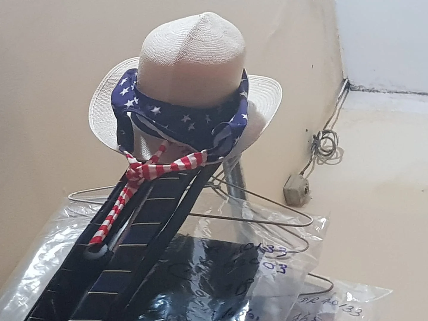 The image displays a collection of items hanging together, likely in a storage or closet area. Central to the image is a white straw hat adorned with a band featuring a star pattern, reminiscent of the American flag. Draped over the hat is a red and white striped scarf, adding a patriotic touch. The items are supported by wire hangers, which are visible beneath a clear plastic cover. The background reveals a beige wall with visible wiring, a plug socket, and a small CCTV camera, indicating an indoor setting. The lighting is soft, suggesting an overhead light source. The scene captures a blend of clothing items with a subtle Americana theme, organized in a functional, though visually striking, manner.