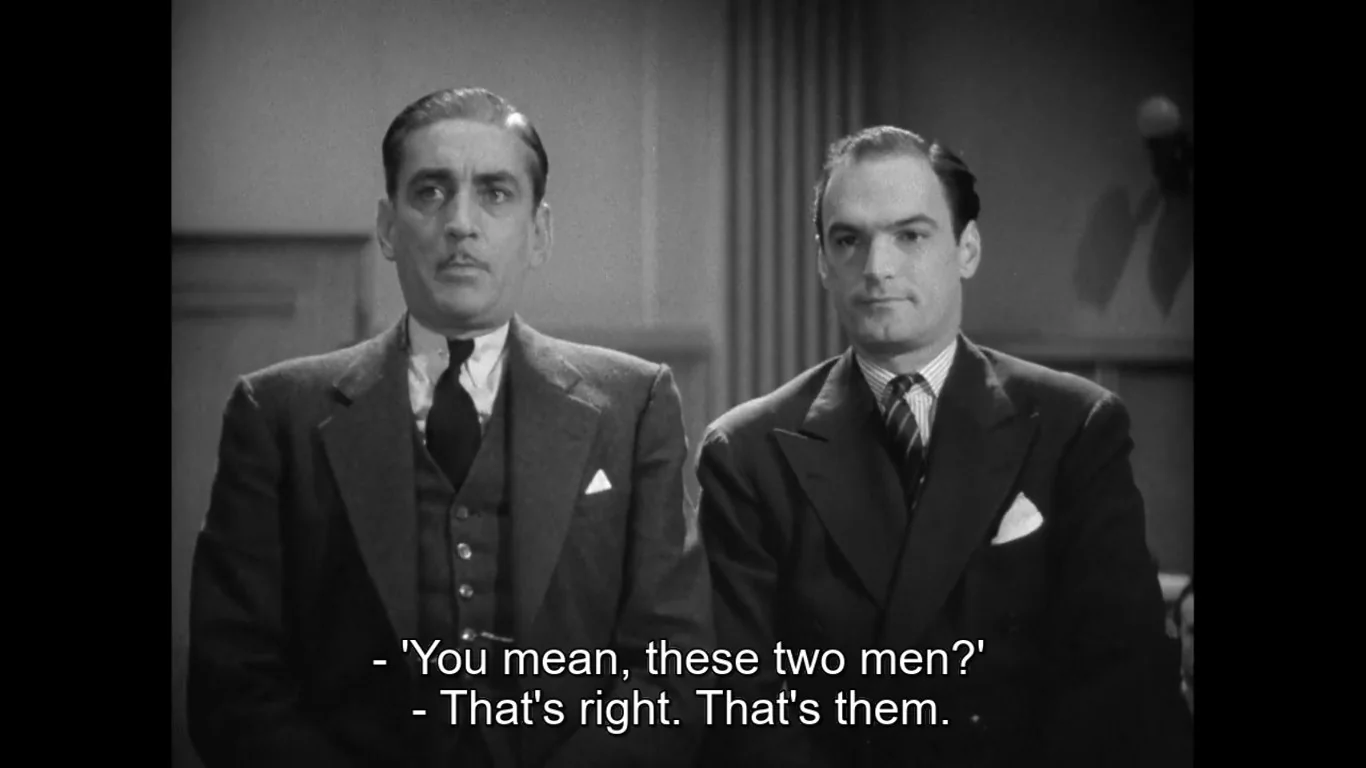 In this classic black-and-white film scene, two men are standing indoors, dressed in formal suits. The man on the left wears a three-piece suit with a dark tie and a neatly folded pocket square, exuding a composed and serious demeanor. His hair is slicked back, adding to his polished appearance. The man on the right is also in a dark suit and tie, with a more relaxed expression. They appear to be in a setting reminiscent of a courtroom or an office, suggested by the plain walls and the sense of formality.
The lighting is soft and even, characteristic of films from the mid-20th century, creating a sense of gravity and focus on the characters. The composition centers on these two men, who stand closely together, suggesting camaraderie or a shared purpose. The subtitles read, "'You mean, these two men?' - That's right. That's them." This dialogue hints at an identification or recognition scene, possibly involving testimony or revelation. The overall mood is serious and dramatic, capturing a moment of significance between the characters.