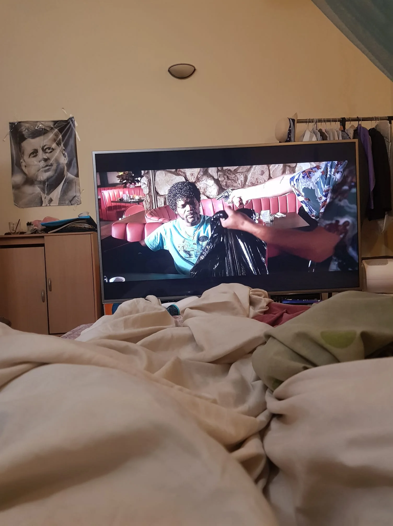 The image shows an indoor setting, likely a bedroom. In the foreground, there are rumpled bed linens suggesting someone is viewing the scene from a bed. In the background, a TV screen is prominently displaying a scene from a movie. The scene features a man seated in a booth, wearing a light blue t-shirt, while someone else hands him a black garbage bag. The setting appears to be a diner with red seating and stone wall decor visible.
To the left, a poster of a historical figure is pinned to the wall above a cabinet with some items on top, including a book or magazine. In the corner, a clothing rack with various garments is visible, adding to the lived-in feel of the room. The lighting is soft and ambient, contributing to a relaxed atmosphere. The room feels personal and cozy, indicative of someone settling in to watch a movie from the comfort of their bed.