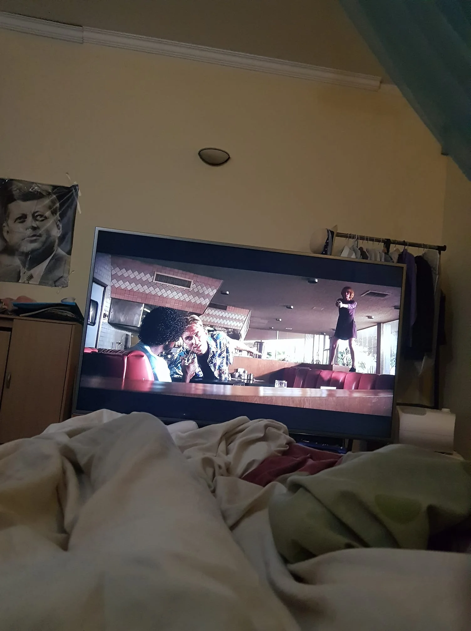 The image depicts a scene from the film "Pulp Fiction" displayed on a television screen in a cozy bedroom setting. The TV shows a tense moment in a diner where one character stands on a table pointing a gun, while another leans in to talk to someone seated. The background features diner decor with red booths and patterned walls, typical of the film's retro style. The room itself is dimly lit, giving it an intimate and relaxed atmosphere. In the foreground, there are unmade bedsheets, suggesting someone is lying down to watch the movie. A closet with hanging clothes is visible to the right of the TV, and there's a partially visible poster on the wall to the left featuring a historical figure. Overall, the setting feels personal and lived-in, creating a contrast between the tranquil environment and the intense movie scene.