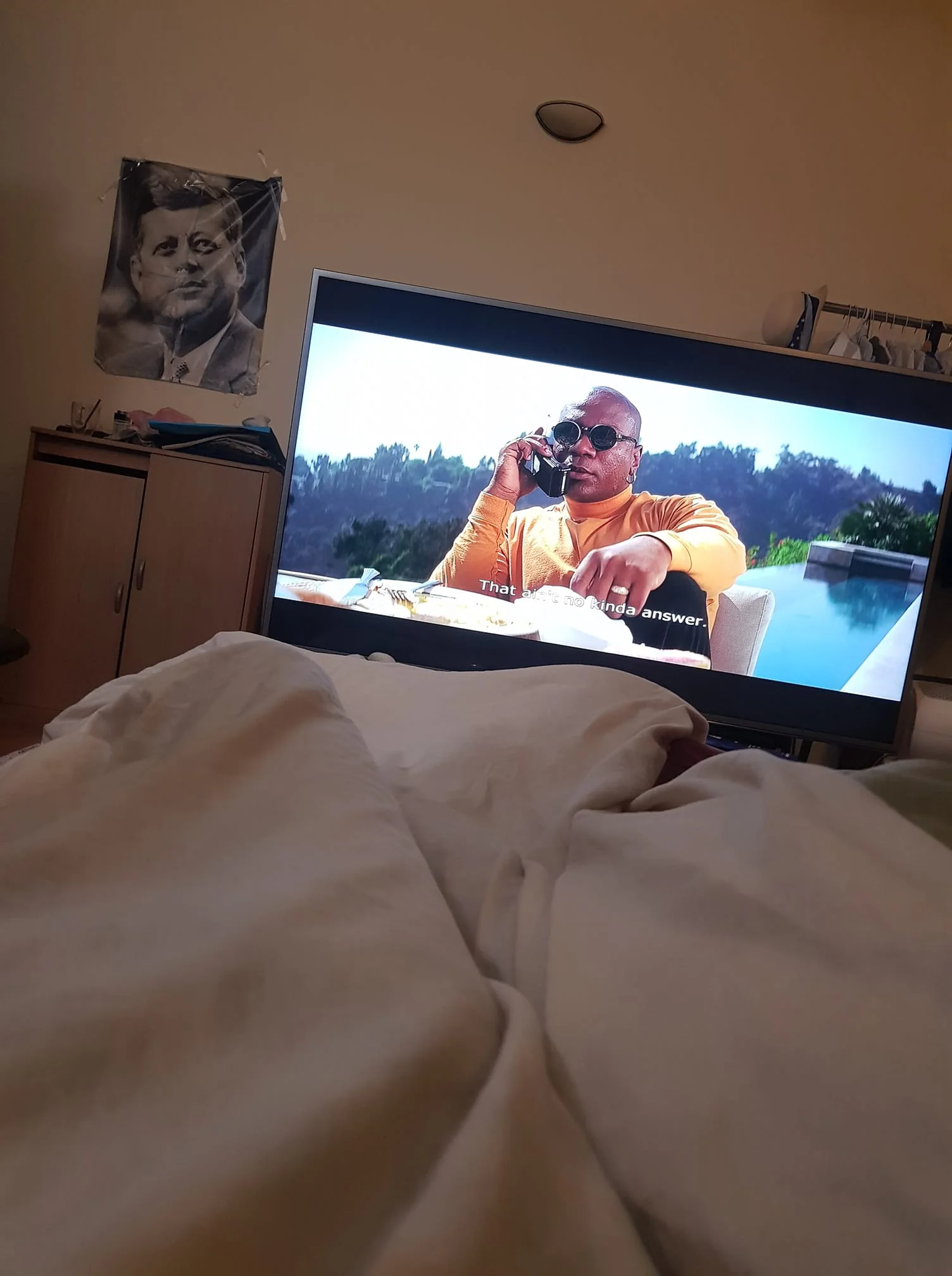 The image shows an indoor scene, likely in a bedroom. In the foreground, a person is lying under a blanket, capturing a cozy and relaxed atmosphere. A television in the room displays a scene with a man wearing a bright orange shirt and sunglasses, speaking on a phone. The setting behind him appears to be outdoors, with a view of trees and a pool, indicating a luxurious location. The subtitle on the screen reads "That's the kinda answer."
In the background, a poster on the wall features an image of Kennedy, adding a historical or political touch to the room’s decor. A wooden cabinet and some clothes on a rack are also visible, providing a lived-in feel. The lighting is soft and natural, suggesting a calm and relaxed environment.