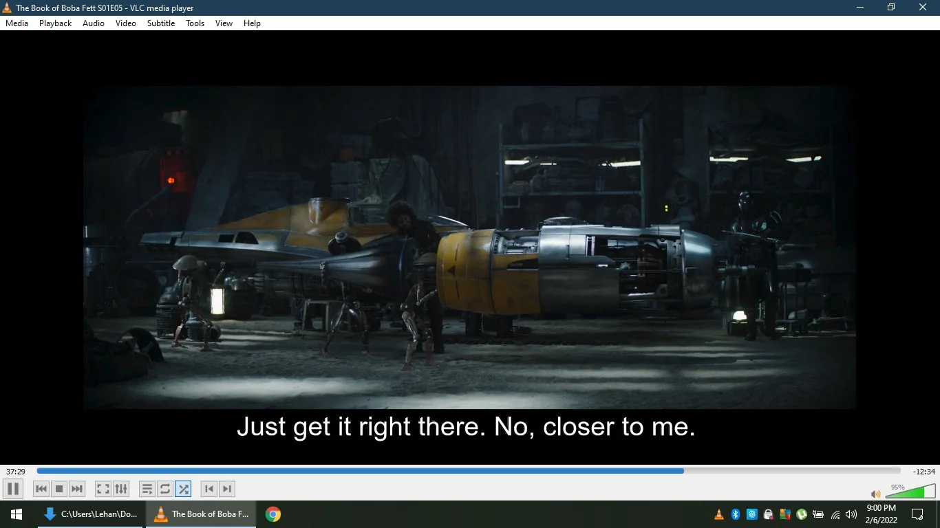 The image is a screenshot from "The Book of Boba Fett," specifically season 1, episode 5, as indicated by the title text in the VLC media player window. The scene takes place in a dimly lit workshop, featuring a partially assembled spacecraft that resembles a Naboo N-1 Starfighter. A character, possibly a mechanic, is working on the ship, assisted by a mechanical droid. Another individual, wearing dark armor typical of the Star Wars universe, stands nearby, observing the process. The lighting is moody, with shadows and focused light highlighting the spacecraft's metallic surfaces. The workshop is cluttered with various tools and equipment in the background. The subtitle reads, "Just get it right there. No, closer to me," indicating a precise task being carried out. The overall atmosphere suggests a scene focused on repair or modification, common in the Star Wars series.
