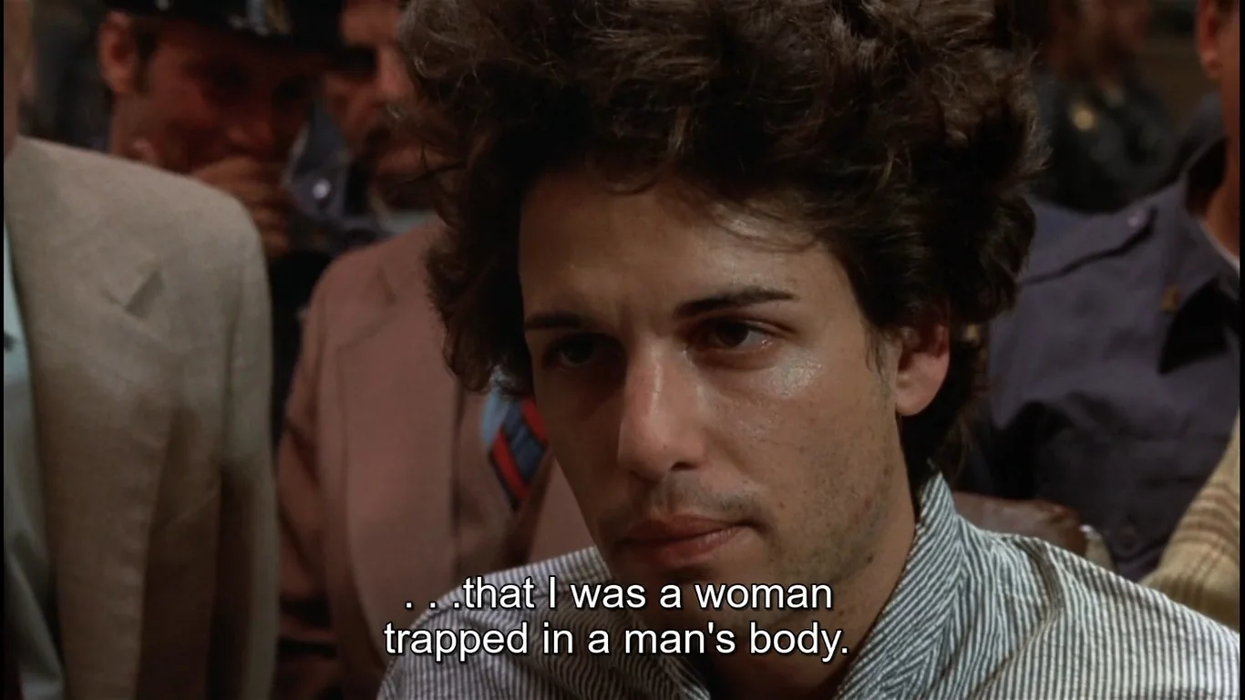 ```
The image is a frame from a movie scene set indoors, depicting a serious and tense atmosphere. The focal point is a person with curly hair, sitting among a group of people, possibly at a hearing or meeting. The person is wearing a striped shirt and has a contemplative expression, suggesting they are involved in a deep or revealing conversation. The background is filled with other people, some in uniforms, indicating a formal or official setting. The lighting is natural, highlighting the central figure's face, enhancing the emotional intensity of the moment. The subtitles at the bottom of the frame read, "...that I was a woman trapped in a man's body," indicating a discussion about identity and personal revelation. The scene captures an intimate and emotional moment, reflecting themes of gender identity and self-disclosure.