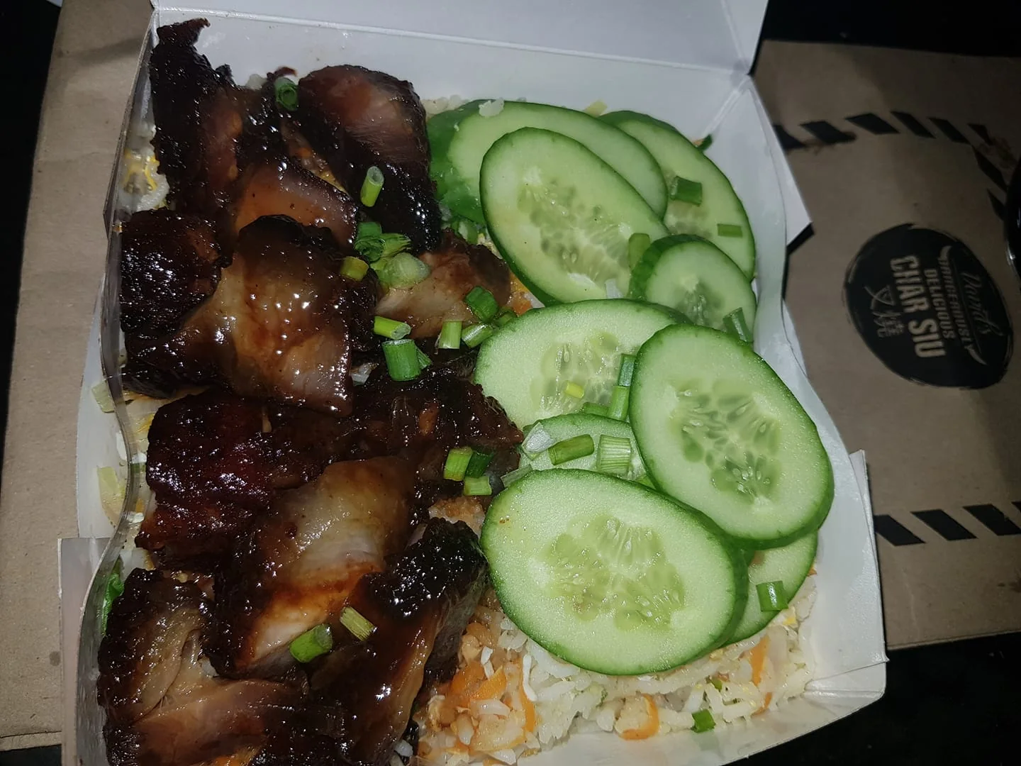 This image showcases a delicious-looking meal that features char siu, a popular Chinese barbecue pork dish. The char siu appears to be rich and glossy, possibly glazed with a sweet and savory sauce, giving it a dark, caramelized appearance. The dish is served in a rectangular takeout container, which is lined with a piece of wax paper or similar material. Accompanying the char siu are thinly sliced cucumbers arranged neatly on one side, adding a fresh and crisp contrast to the meal. Below, there seems to be a base layer of fried rice, speckled with bits of vegetables and garnished with sliced green onions. The setting suggests an indoor environment, possibly a restaurant or home, and the lighting is moderate, focusing on the meal's vibrancy. A part of the packaging visible in the background contains the text "Deliciously Char Siu," hinting at the brand or restaurant specializing in this dish. The composition highlights the colorful and appetizing nature of the meal, making it the focal point of the image.
