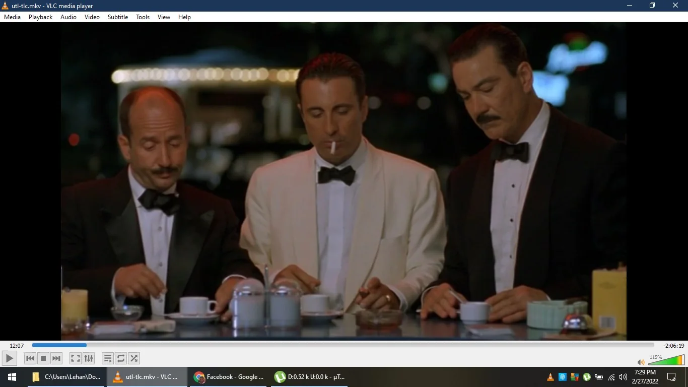 The image is a screenshot from VLC media player showing a scene with three men dressed formally in tuxedos, suggesting a setting of sophistication or a significant event. The man in the center, identified by the hint as Andy Garcia, is wearing a white tuxedo jacket with a black bow tie and is holding a cigarette. His expression is contemplative, as he looks down at the table. The two men on either side of him wear black tuxedos and appear to be focused on what they are doing, possibly preparing or stirring their drinks. The setting resembles a diner or bar counter, with cups, a sugar dispenser, and other typical items present. The lighting is dim, creating a nighttime ambiance. The VLC media player's interface is visible, showing the file path "C:\\Users\\Lehan\\Do..." and the video file name "utl-tlc.mkv."
