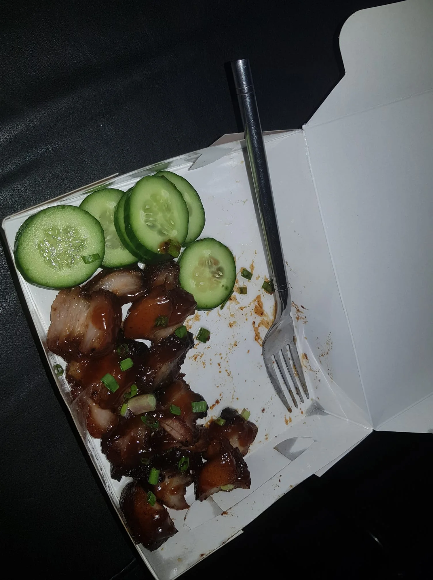 The image shows an open takeout box containing slices of glazed meat garnished with chopped green onions. Next to the meat are several cucumber slices arranged neatly. A metal fork is also present in the box, indicating it may have been used by someone to eat the meal. The inside of the box has some sauce stains, suggesting the meal was recently consumed. The lighting is direct, illuminating the food and creating a focus on its glossy texture and fresh appearance. The setting appears to be indoors, possibly on a dark surface like a table or seat. The overall mood of the image is casual, suggesting a quick meal or snack. The use of takeout packaging indicates convenience and portability.