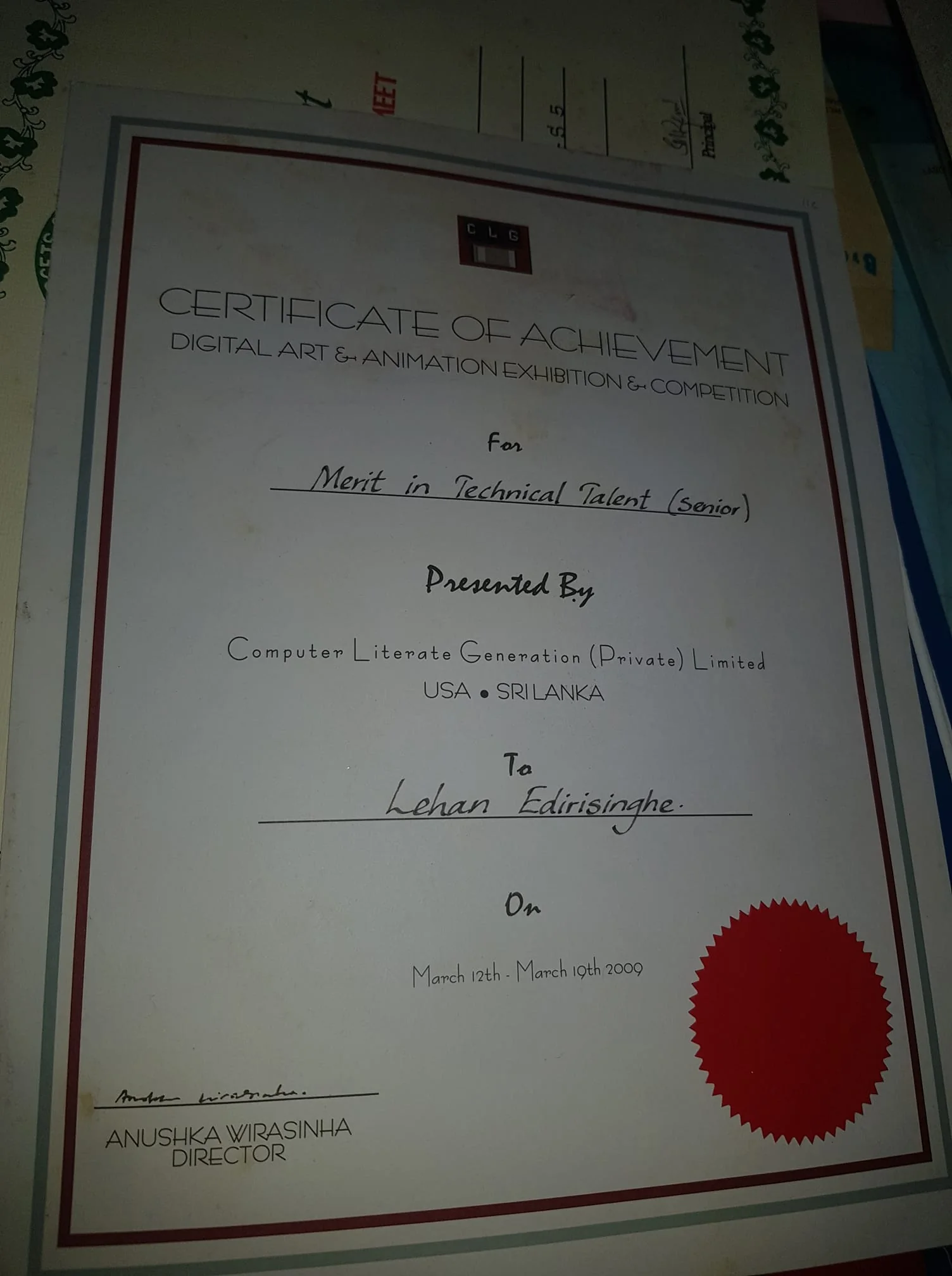 The image shows a Certificate of Achievement awarded for "Merit in Technical Talent (Senior)" in a Digital Art & Animation Exhibition & Competition. The certificate is presented by Computer Literate Generation (Private) Limited, located in both the USA and Sri Lanka. It is awarded to someone named Lehan Edirisinghe and is dated from March 12th to March 19th, 2009. The certificate is signed by Anushka Wirasinha, who is identified as the Director.
The document is designed with a formal layout, featuring a red and silver border. A red seal is placed on the bottom right of the certificate, adding a touch of authenticity. The text is printed in different fonts, with important details like the recipient's name and the director’s signature handwritten. The certificate is slightly tilted in the image, placed on a table alongside other documents, indicating it might be part of a collection. The lighting is dim, casting soft shadows on the paper, suggesting an indoor setting.