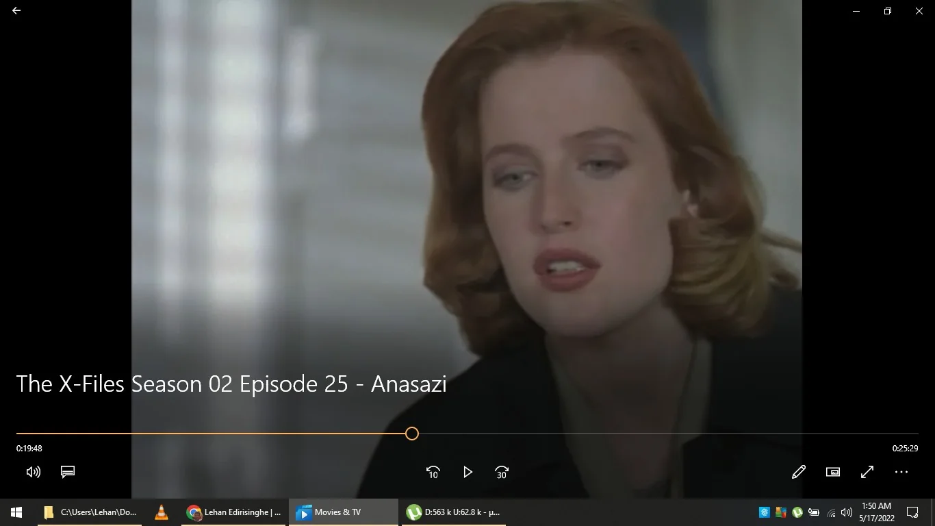 The image is a frame from a video playing on the Movies & TV app. The title text indicates it is from "The X-Files Season 02 Episode 25 - Anasazi". The scene features a close-up of a person with red hair, looking contemplative or engaged in conversation. The lighting is soft, and the background appears blurred, suggesting an indoor setting. The video playback controls are visible at the bottom, showing a timestamp of 0:19:48 out of 0:25:29. The interface has standard playback options like play, rewind, and volume control. The desktop taskbar at the bottom of the image shows system icons and a timestamp of 1:50 AM on 5/17/2022. The overall mood of the scene seems serious, matching the investigative and mysterious themes typical of "The X-Files."