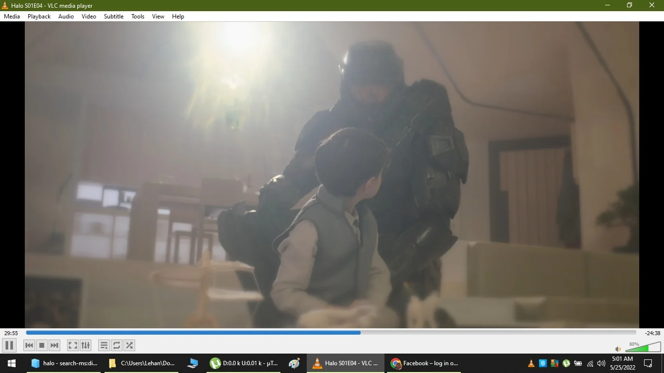 The image is a screenshot from a computer screen showing a scene from the series "Halo," specifically episode 4 of season 1, as indicated by the title "Halo S01E04 - VLC media player." The scene is displayed in VLC media player, which is visible on the screen with its typical control interface at the bottom.
In the scene, a young boy is seen interacting with a figure dressed in futuristic, armored attire typical of the Halo universe. The setting appears to be indoors, possibly in a living room or similar space, with soft lighting coming from a window or light source in the background, creating a bright halo effect around the characters. The armored figure, likely a super-soldier from the series, is seated and appears to be in conversation with the boy, who is looking up at him. The boy is wearing a light-colored shirt and vest, contributing to a calm and neutral color palette for the scene.
The computer's taskbar shows that it is 5:01 AM on May 25, 2022. There are various other icons and windows visible, indicating typical computer use. The presence of the Halo series suggests a science fiction theme with elements of futuristic technology and military influence.