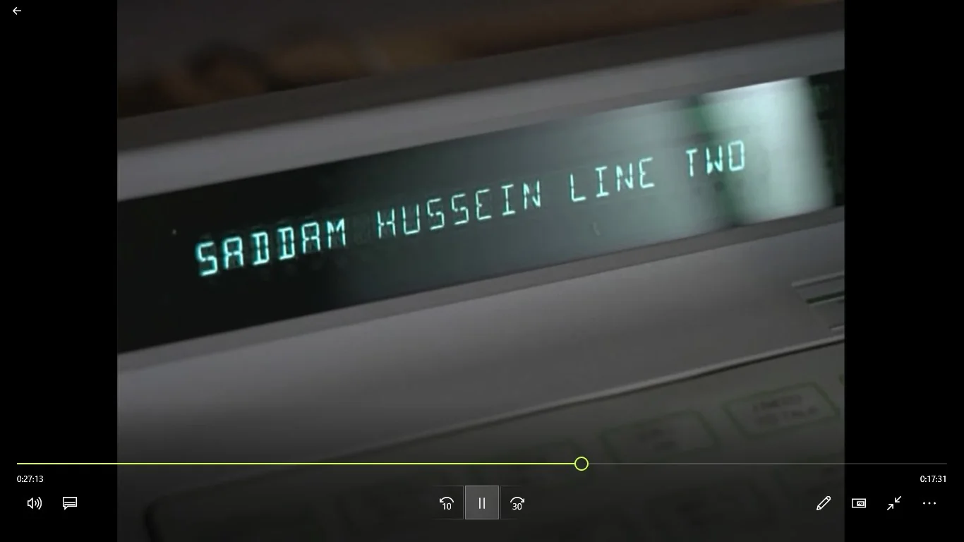 The image features a close-up view of an electronic display screen, likely a phone or communication device, in an indoor setting. The screen prominently displays the text "SADDAM HUSSEIN LINE TWO" in bright, greenish-blue lettering against a dark background. This suggests an incoming call or message from someone labeled as Saddam Hussein, indicating a fictional or humorous context. The device itself appears modern, with visible buttons on the lower right corner, possibly used for handling communication functions. The lighting is soft, casting a slight reflection on the screen, contributing to a professional and somewhat tense atmosphere. The scene might be from a film, television show, or other media where the character or entity named is part of the narrative.