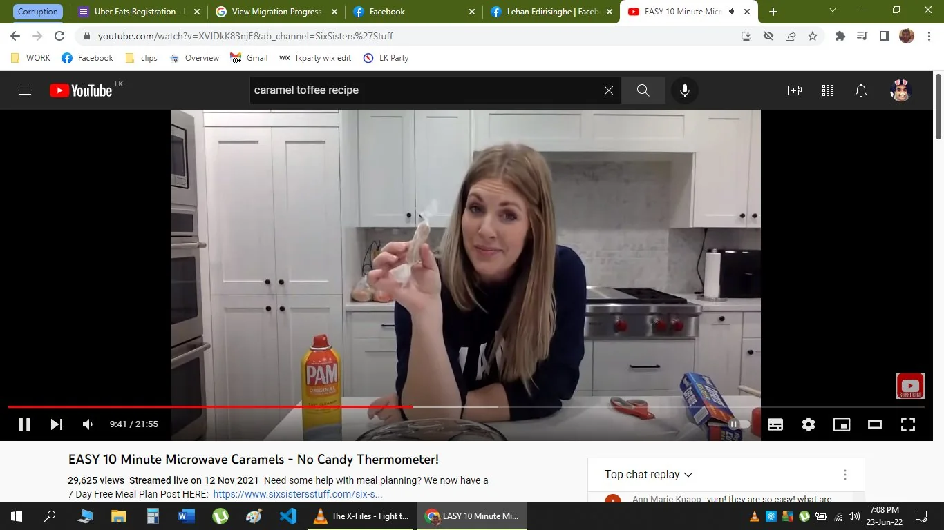 The screenshot shows a YouTube video titled "EASY 10 Minute Microwave Caramels - No Candy Thermometer!" from the channel "SixSisters'Stuff." In the video, a woman is standing in a well-lit, modern kitchen. She is wearing a dark shirt and has long, light-colored hair. She is holding a small item in her hand, possibly related to the caramel recipe she is demonstrating. The background features white cabinets, a stovetop with red knobs, and a countertop with various items, including a can of PAM spray and a box of foil. The mood appears instructional and friendly, as she engages with the audience. The scene is set for a cooking tutorial, focusing on a quick and easy method to make caramels using a microwave, without needing a candy thermometer. The video is part of a series aimed at providing simple and accessible cooking solutions.