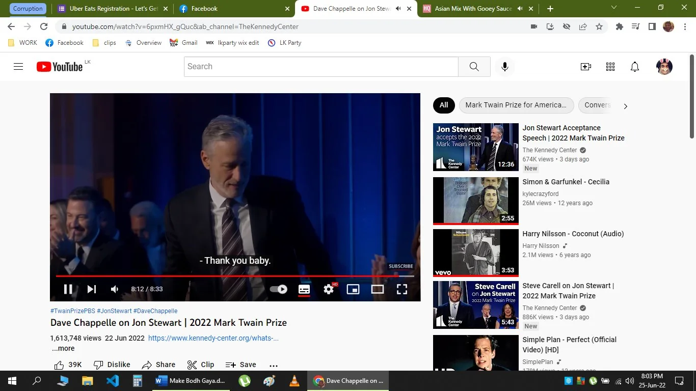 The image is a screenshot from a YouTube video titled "Dave Chappelle on Jon Stewart | 2022 Mark Twain Prize," hosted on The Kennedy Center's channel. The scene captures an indoor event, likely an award ceremony, since it involves the Mark Twain Prize. The video thumbnail shows a person on stage, presumably Jon Stewart, as suggested by the hint. The individual is dressed in a formal suit, standing in front of a dark blue curtain backdrop, suggesting a formal and elegant setting. The atmosphere appears celebratory, with an audience visible in the background, including people applauding. The text "Thank you baby" is part of the captions, indicating a moment of gratitude or affection. The video seems to focus on Jon Stewart, who is a central figure in the event. The YouTube interface is visible, with various video recommendations related to Jon Stewart and other similar topics on the right sidebar.