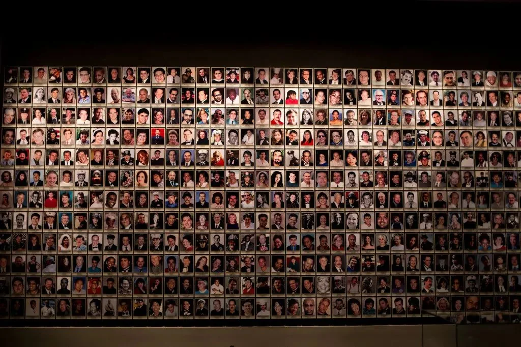 This image captures a vast wall filled with numerous small portraits, arranged in a dense grid pattern. Each portrait is a photograph of an individual, showcasing a diverse range of people, likely representing a memorial or tribute display. The lighting in the room is subdued, casting a warm glow that highlights the individual faces, each captured in unique expressions ranging from solemn to serene. The photographs are uniformly sized, creating a cohesive and structured visual presentation.
The setting appears to be indoors, possibly in a gallery or museum, where visitors might spend time reflecting on the images. The photographs vary in color, with some in vivid detail and others in black and white, indicating a possible historical or commemorative aspect. The overall mood is somber and respectful, suggesting the significance of the individuals represented. While the exact purpose or context of the exhibit is not specified, the sheer number of portraits implies a large-scale memorial, honoring the memory of those pictured.