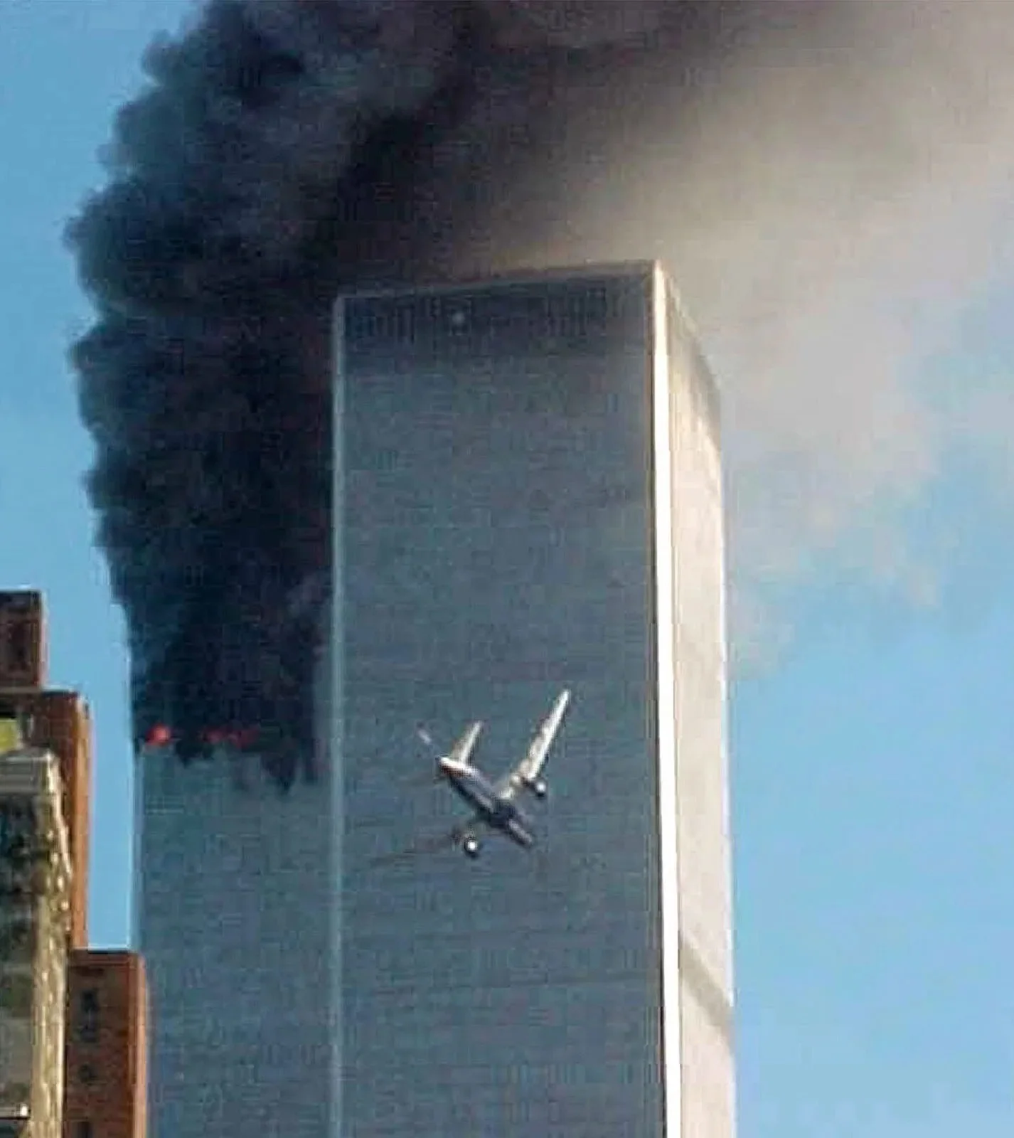 The image captures a critical moment from the September 11, 2001, attacks in New York City. A commercial airplane is seen approaching the South Tower of the World Trade Center, with the North Tower already engulfed in thick, black smoke. The photograph is shot in daylight, with clear blue skies contrasting with the dark smoke emanating from the building. The plane is angled slightly towards the tower, highlighting the impending collision. The South Tower stands prominently to the right, while the North Tower, partially obscured by smoke, is to the left. The scene is tense, capturing the catastrophic events as they unfold. The urban setting is evident, with hints of other buildings in the lower left, emphasizing the cityscape where this tragedy occurred. The image conveys a sense of urgency and foreboding, marking a pivotal moment in modern history.