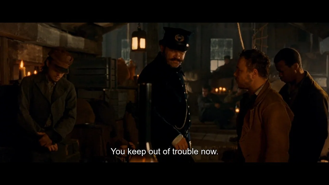 ```
The image shows a dimly lit indoor scene, likely from a period drama or historical film. The setting appears to be a rustic interior with wooden beams, crates stacked against the walls, and flickering candlelight creating a moody atmosphere. At the center is a man in a dark blue uniform and cap, suggestive of 19th-century law enforcement, with a noticeable mustache. He is in conversation with another man dressed in a brown jacket, facing him. The caption, "You keep out of trouble now," suggests a warning or advice being given. Two other men are present, one leaning against the wall with his head down, and another standing nearby, both adding to the sense of a tense or serious exchange. The lighting is low, emphasizing the shadowy, clandestine feeling of the setting.