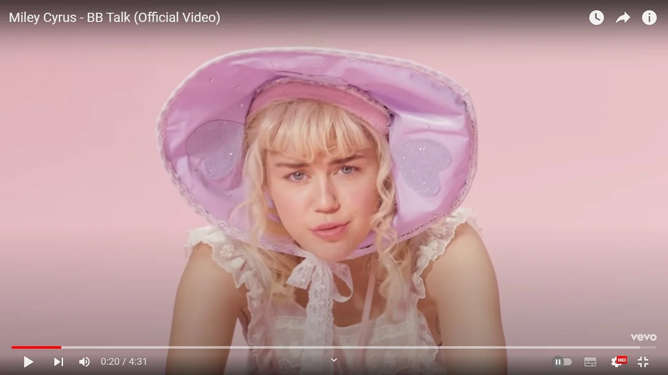 Miley Cyrus appears in a distinctive style in this frame from the "BB Talk" official music video. She is wearing a large, pastel pink bonnet adorned with heart-shaped decorations and lace trim. Her hair is styled in loose curls with bangs, and she wears a light pink headband beneath the bonnet. Her outfit is a delicate, lace-trimmed garment that resembles a vintage baby dress, contributing to a playful, whimsical aesthetic. The background is a soft, uniform pink, complementing the outfit's color scheme and creating a cohesive visual theme. The lighting is bright and evenly distributed, highlighting her facial expression, which is both inquisitive and slightly pouty, enhancing the playful mood of the scene. The video is published by Vevo on MileyCyrusVEVO's YouTube channel, and the frame captures the unique, stylized approach of the "BB Talk" music video.