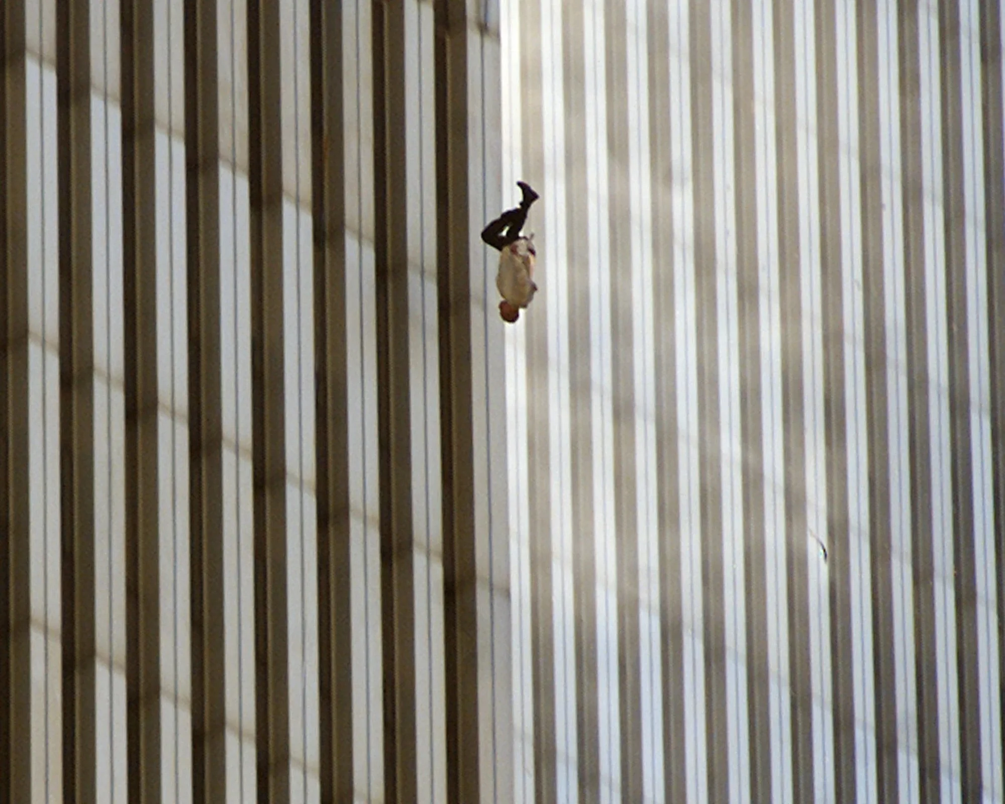 The image captures a harrowing moment during the 9/11 attacks, showing an individual falling from one of the Twin Towers. The person is upside down, wearing a white shirt and dark pants. The backdrop is the façade of the building, characterized by its vertical lines and reflective windows. The lighting casts shadows across the surface, emphasizing the tower's structural design. This photograph conveys a sense of tragedy and urgency, encapsulating the chaos and desperation of that day. The lack of additional elements in the image, such as other people or objects, focuses attention entirely on the falling individual, highlighting the human impact of the event. The image is somber and poignant, serving as a powerful reminder of the events of September 11, 2001.