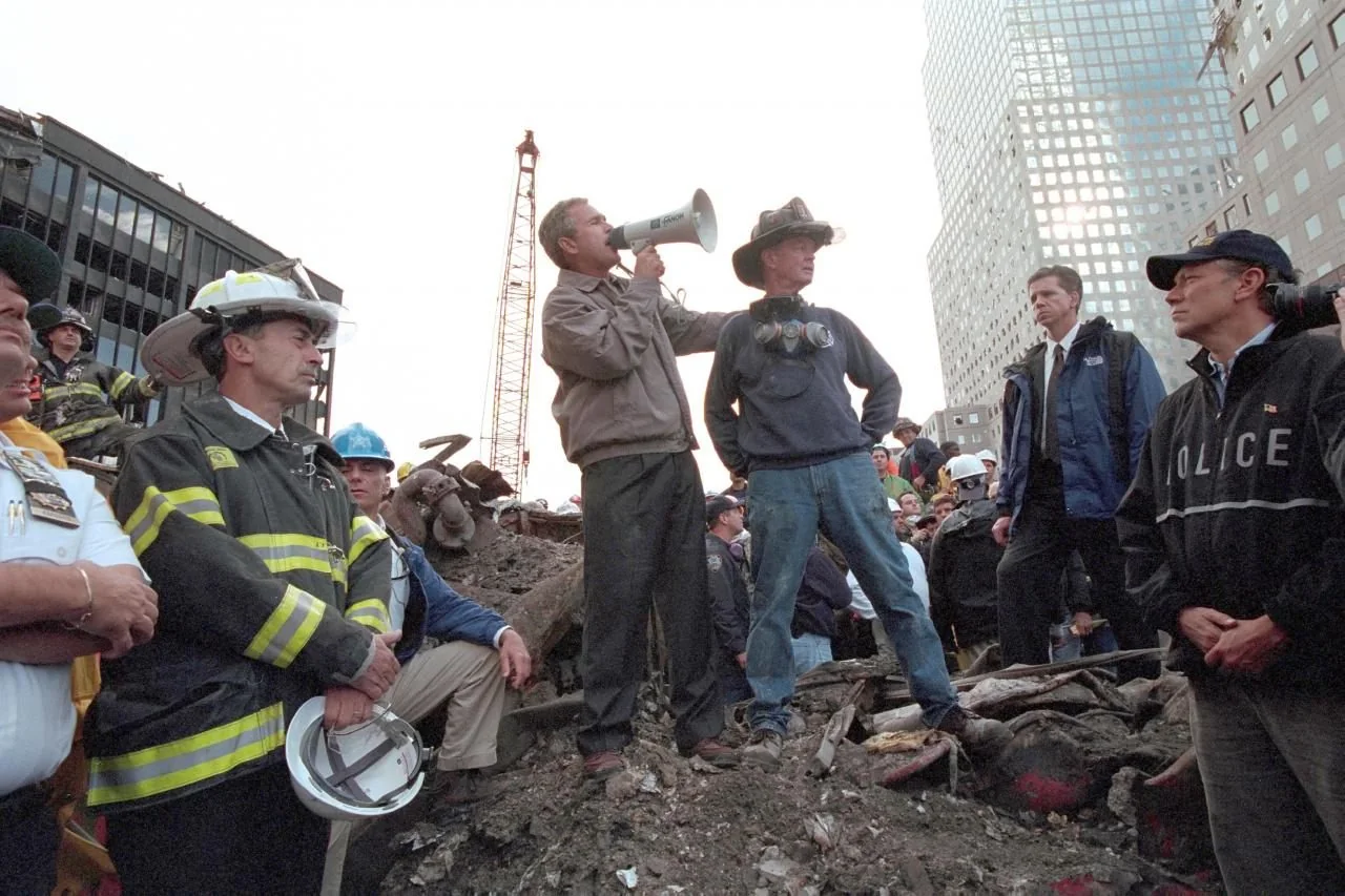 In this image, George W. Bush is seen standing amidst the debris of a disaster scene, using a megaphone to address a crowd. The setting appears to be an outdoor rescue site, likely following a significant event requiring emergency response. George W. Bush is positioned on a pile of rubble, surrounded by rescue workers and officials, some wearing helmets and protective gear. The atmosphere is serious and somber, reflecting the gravity of the situation. In the background, partially damaged buildings and a construction crane are visible, adding to the chaotic scene. The lighting is natural, indicating it is daytime, and the composition centers on George W. Bush, emphasizing his role in the moment. The image captures a moment of leadership and solidarity in the wake of a crisis.