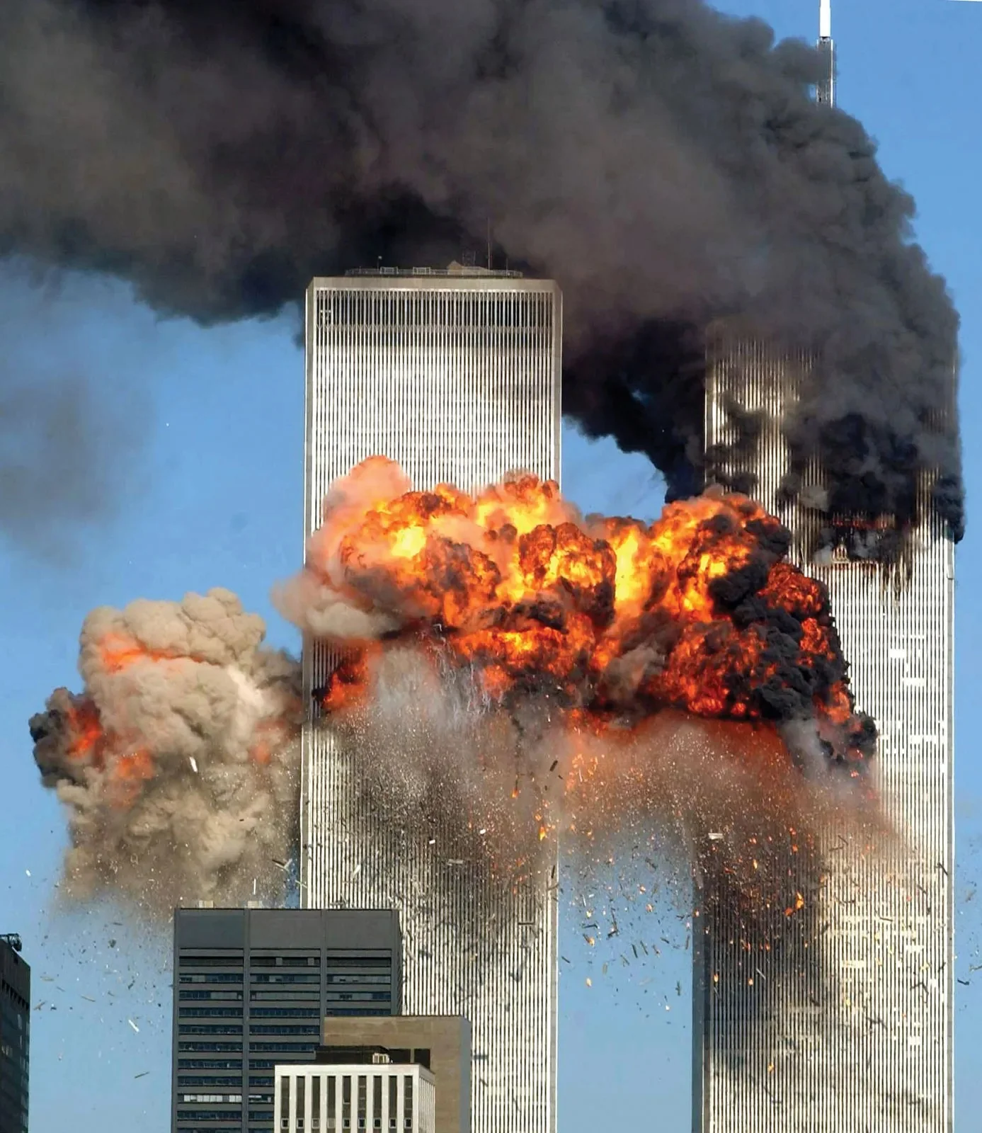 This image captures a pivotal and tragic moment during the September 11, 2001, terrorist attacks on the World Trade Center in New York City. The photograph shows a massive explosion in the South Tower, with flames and thick black smoke billowing outwards. The bright orange and red flames contrast starkly against the blue sky, creating a visually striking yet devastating scene. Debris is visible flying out from the impact site, adding to the chaotic energy of the moment. The North Tower, already burning, stands adjacent, with dark smoke obscuring its upper section. Below, smaller buildings are partially visible, dwarfed by the towering structures. The image conveys a sense of destruction and urgency, encapsulating one of the darkest days in modern history.