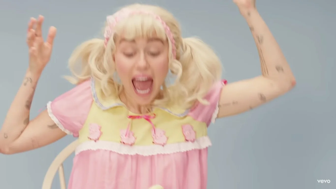 Miley Cyrus is featured in this video frame, which captures an energetic and playful scene. She is wearing a pink and yellow dress adorned with decorative elements, giving a childlike or whimsical appearance. Her hair is styled in blonde pigtails with a pink headband. Miley's facial expression is animated, as she appears to be mid-shout or scream, with her mouth wide open and eyes expressive. Her arms are raised in an exaggerated gesture, contributing to the dynamic and lively mood of the scene.
The background is a simple light blue, which highlights her brightly colored outfit and her energetic posture. Her arms display tattoos, adding a personal touch to her appearance. The lighting is bright and even, emphasizing the vivid colors and the playful atmosphere of the video. The branding in the corner identifies the channel as "vevo," suggesting this is a professional music video frame. There is no additional text or dialogue visible in this frame.