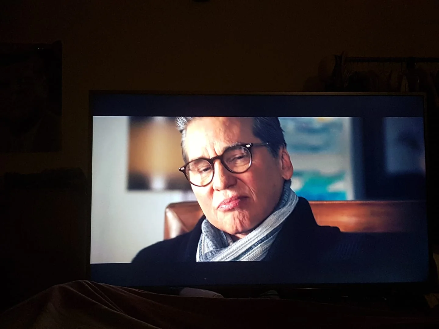 The image is a video frame depicting an older person wearing glasses, a dark jacket, and a light-colored scarf. The scene is set indoors, with a softly blurred background featuring abstract art with blue and beige tones, suggesting a professional or artistic setting. The individual's expression is contemplative, with slightly pursed lips, giving an impression of deep thought or reflection. The lighting is warm and focused on the person, highlighting their features against the darker background. The composition centers the individual, capturing them from the shoulders up, while a wooden chair is partially visible. The mood conveyed is introspective, possibly indicating an interview or personal conversation. The frame does not contain any visible text or branding, keeping the focus on the subject's expression and attire.