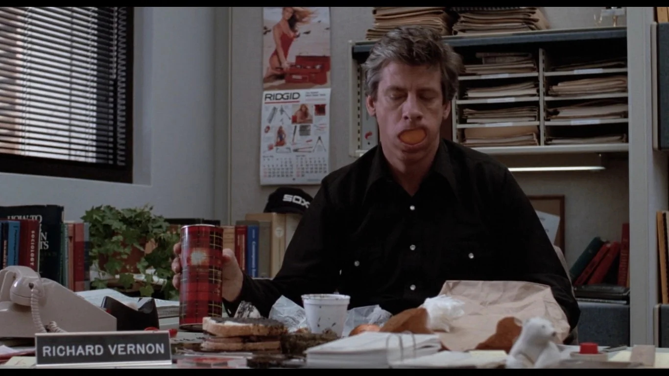 In this scene from "The Breakfast Club," the character Richard Vernon is depicted in his office. He is sitting at a cluttered desk, surrounded by various items including a phone, a plant, and a plaid thermos. Richard Vernon, dressed in a black shirt, has a notable expression with an orange in his mouth, suggesting a humorous or exasperated mood. Behind him, shelves are filled with stacks of folders and papers, indicating a busy or disorganized workspace. The wall features a calendar with the brand "RIDGID" visible, adding to the realistic office environment. The lighting is bright, highlighting the details on the desk and the room's content, such as a nameplate displaying "RICHARD VERNON." A poster with a model in a red outfit is seen in the background, adding a touch of character to the office setting. The overall mood is comedic, fitting the film’s tone.