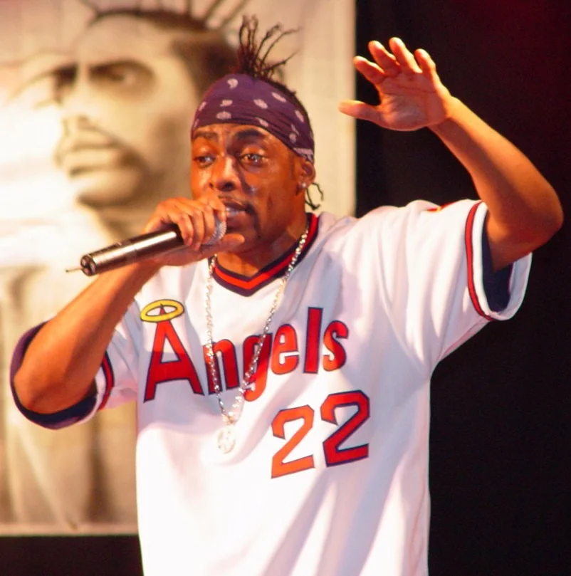 The image captures a vibrant live performance. The performer is wearing an "Angels" baseball jersey with the number 22, and a bandana with a pattern tied around their head. They are holding a microphone, mid-performance, with one hand raised. The expression is intense, indicating a high-energy moment in the performance. In the background, there is a large black-and-white poster featuring an image of a person. The lighting is warm, highlighting the performer against the darker backdrop. The overall scene suggests a lively atmosphere typical of a concert or live music event.