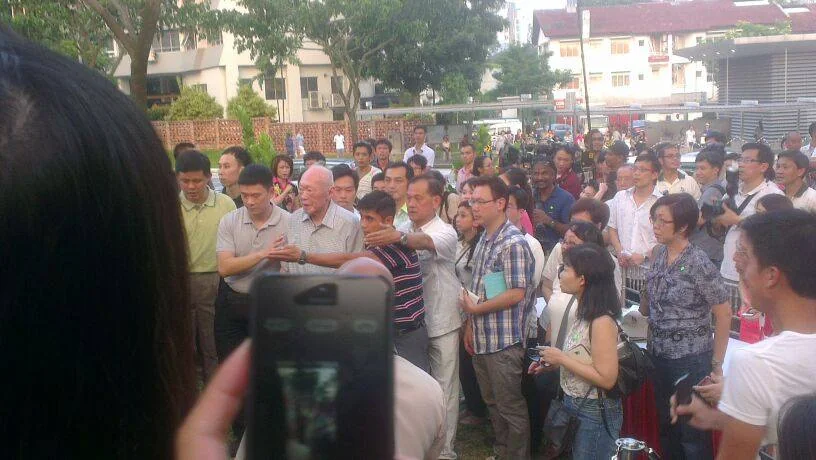The image captures an outdoor public gathering featuring a group of people in what appears to be an informal event or gathering. In the center, an elderly man, identified by the hint as Lee Kuan Yew, is surrounded by individuals who seem to be engaging with him. He has white hair and is wearing a light-colored, short-sleeve shirt. The surrounding crowd consists of people of various ages, some holding cameras and smartphones, suggesting an interest in capturing the moment. Many attendees are casually dressed in shirts and jeans, reflecting a relaxed atmosphere. The background shows trees and a building, indicating the event is taking place in an open, urban area. It is daytime, as evidenced by the natural light illuminating the scene. A person's hand holding a smartphone is visible in the foreground, taking a picture or video of the event. The overall mood is one of interest and engagement from the crowd towards the central figure.