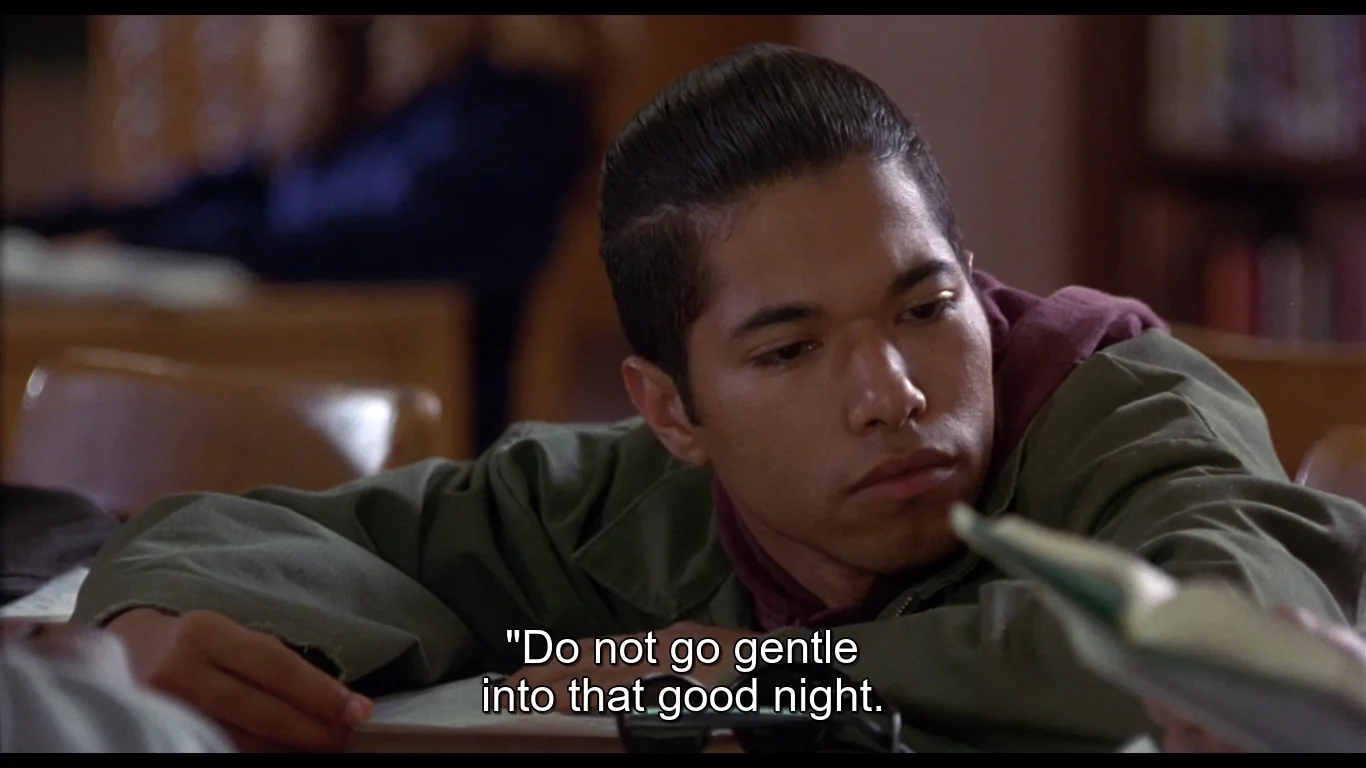 The image is a scene from a video frame set in what appears to be a library or classroom. It features a young man leaning on a table, looking contemplative and somewhat pensive. He is dressed in a green jacket over a maroon shirt, with his head resting on his hand, suggesting a moment of introspection or fatigue. The background is softly blurred, indicating a focus on the individual in the foreground. Books and shelves are visible, reinforcing the library or educational setting. The lighting is soft and natural, giving the scene a calm, subdued atmosphere. The subtitle at the bottom of the frame reads, "Do not go gentle into that good night," a famous line from a poem by Dylan Thomas, suggesting themes of resistance and defiance. The overall mood of the scene is thoughtful and introspective.
