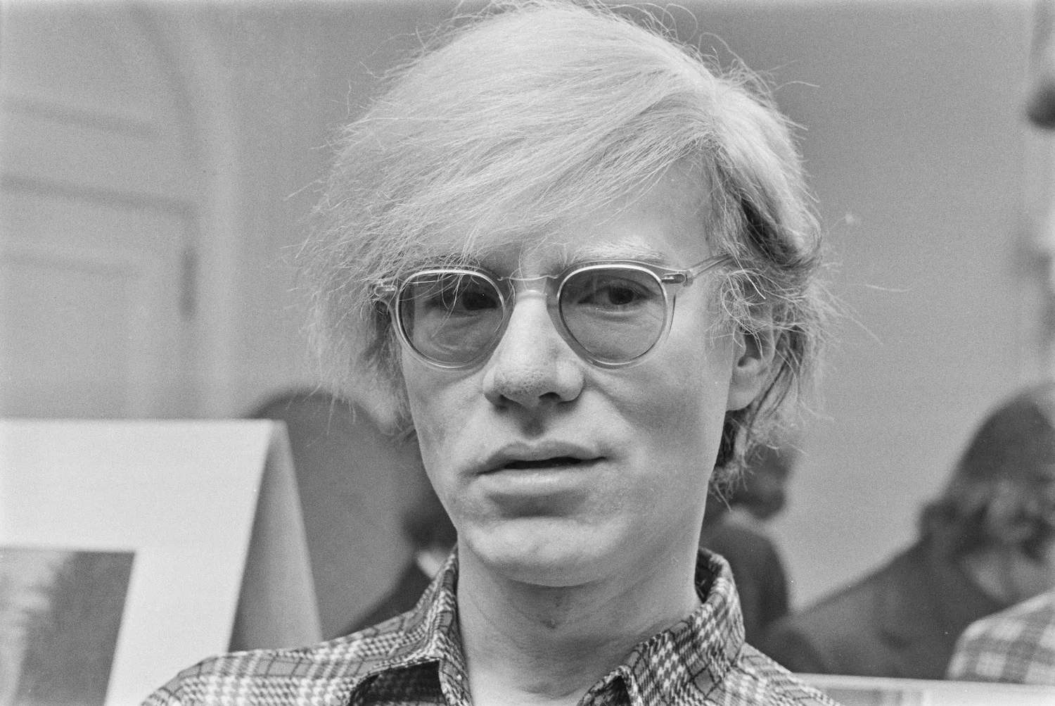 This black and white photograph features Andy Warhol, a prominent figure known for his influence in the visual art movement of pop art. The image is a close-up portrait capturing Warhol indoors, possibly at an art exhibition given the blurred background and presence of artwork. Warhol is wearing a plaid shirt and clear-framed glasses, giving a casual and thoughtful appearance. His signature wispy, light-colored hair is slightly tousled, adding to the iconic look he is known for. The lighting is soft, emphasizing the textures of his hair and clothing, contributing to the overall neutral and contemplative mood of the photograph. There's a focus on his expression, which is calm and slightly introspective, suggesting a moment of pause amidst a busy setting.