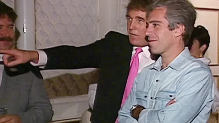 The image depicts a social gathering in an indoor setting, where two individuals, identified by the hint as Trump and Epstein, are engaged in conversation. Trump, on the left, is wearing a black suit with a white shirt and a bright pink tie. He is gesturing with his right hand, pointing towards something out of the frame, indicating that he might be explaining or discussing a point. Epstein, on the right, is dressed more casually in a light denim shirt, with his arms crossed, suggesting a relaxed demeanor. The background shows other attendees and typical party elements like drinks and decorations. The lighting is warm, typical of an indoor evening event, creating a soft ambiance. The mood appears informal and conversational. The presence of other partially visible people in the background indicates a lively atmosphere.