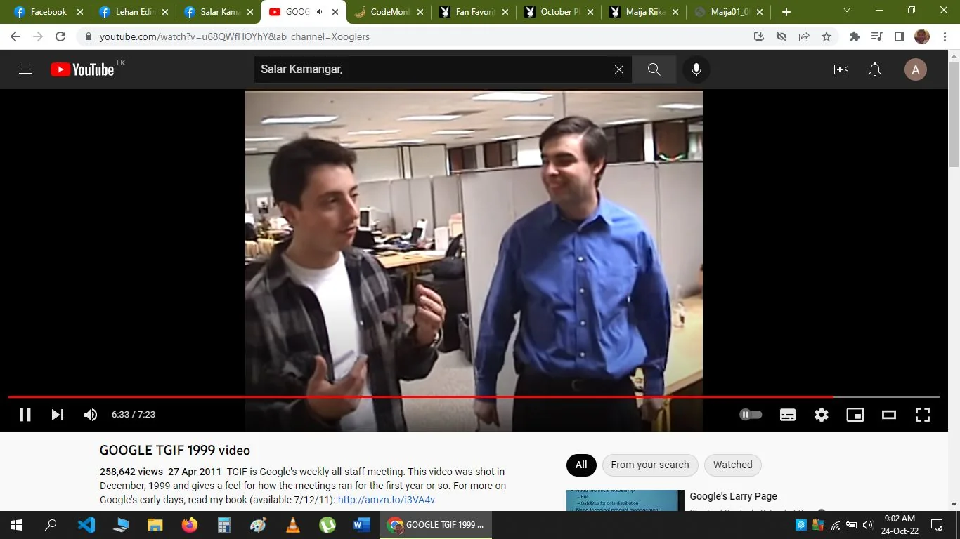 The image is a screenshot from a YouTube video titled "GOOGLE TGIF 1999 video," shared on the "Xooglers" channel. The video features Sergey Brin and Larry Page, co-founders of Google, engaged in conversation in what appears to be an office setting. Sergey Brin, on the left, is wearing a casual plaid shirt over a white t-shirt, gesturing with his hands as he speaks. Larry Page, on the right, is dressed in a blue collared shirt, smiling warmly at Sergey. The setting is an open-plan office with cubicles visible in the background, suggesting a casual and collaborative work environment typical of late 1990s tech companies. The lighting is bright, and the atmosphere appears relaxed and informal. The on-screen text on the YouTube interface includes key details about the video's context, indicating that it was shot during a weekly all-staff meeting, known as TGIF, at Google in December 1999. The office equipment and decor reflect the period, adding to the nostalgic feel of the video.