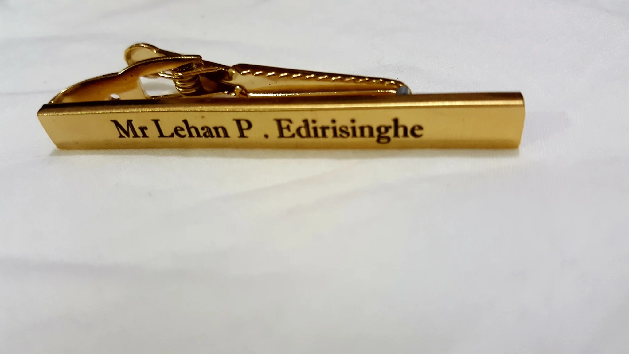 The image showcases a close-up of a gold tie clip with a white background. The tie clip has a polished finish and features engraved text that reads "Mr Lehan P. Edirisinghe." The text is centered and appears in a classic serif font, providing a formal appearance. The clip is slightly angled, casting a soft shadow on the surface. The gold color of the clip contrasts with the plain white background, highlighting its elegant design. The spring mechanism of the clip is partially visible, indicating functionality. The setting suggests a focus on the object, with sufficient lighting highlighting its metallic sheen. No other objects or details are visible in the image, keeping the emphasis solely on the tie clip.