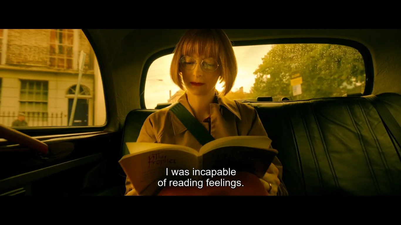 This image depicts a scene inside a car with a woman reading a book titled "The Prophet." The lighting is warm and casts an orange hue, suggesting it might be either early morning or late afternoon. The woman has short brown hair and is wearing glasses, which add an intellectual touch to her appearance. She is seated in the backseat of the car, and her seatbelt is visibly fastened across her chest. The background outside the car window is slightly blurred, indicating motion, with trees and a building passing by. The atmosphere inside the car feels introspective, enhanced by the subtitle "I was incapable of reading feelings," which suggests a theme of emotional disconnect or introspection. The woman appears focused and absorbed in her reading, contributing to a quiet, contemplative mood.